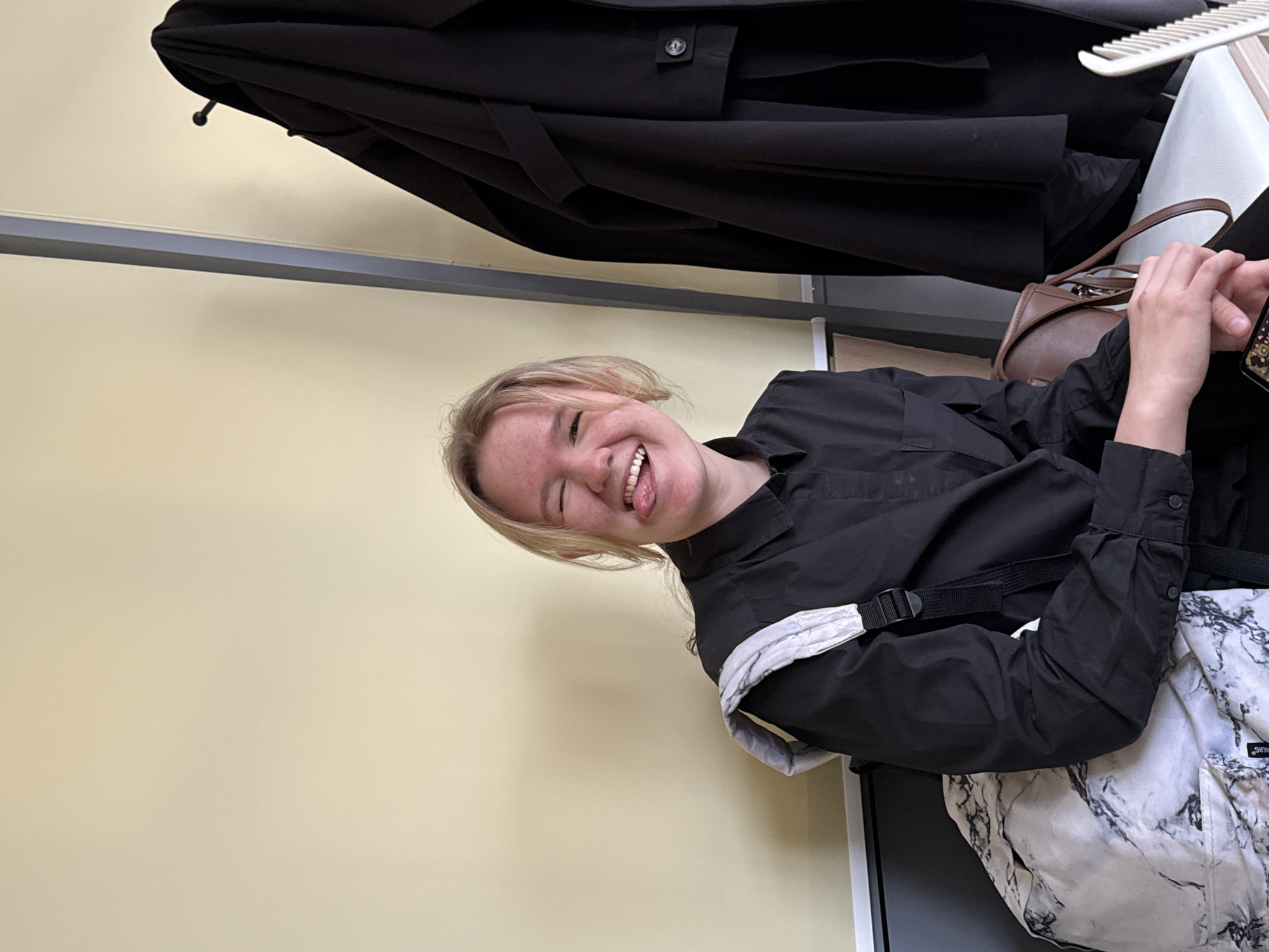 A young woman with blonde hair is sitting in front of a pale yellow wall. She is wearing a black collared shirt and a backpack with a white and gray marble pattern. She is sticking her tongue out and smiling. Above her, a black coat is hanging on a hook. A white comb rests on a white surface next to a brown purse. The location is potentially Chaikovsky, Russia.