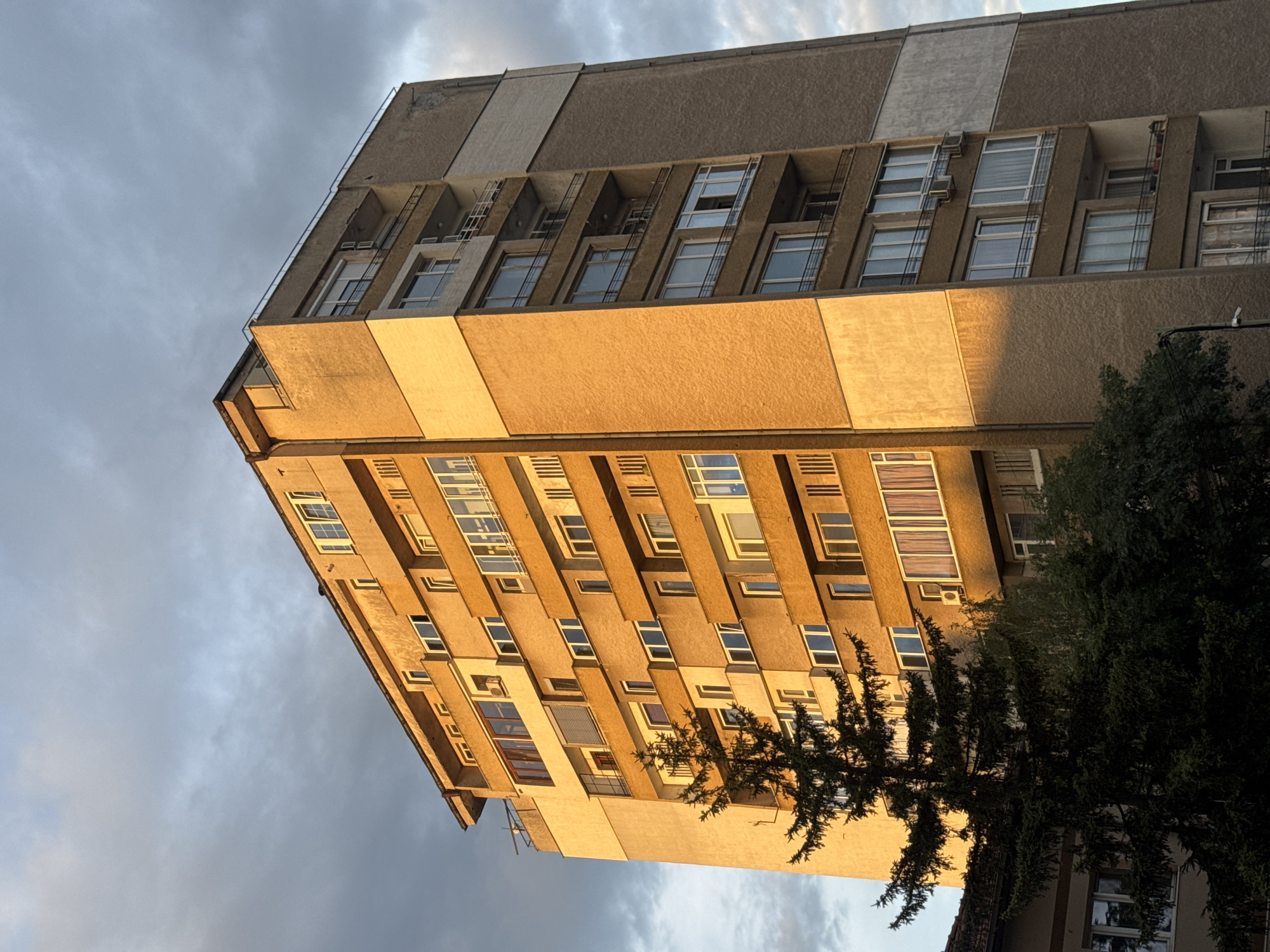 This is a vertical, eye-level, medium shot of a residential apartment building bathed in the warm, golden light of sunset, casting long shadows across its facade. The building, characterized by its brutalist architectural style, features multiple floors with balconies and numerous windows. The exterior walls are a textured brown, interspersed with lighter cream-colored rectangular panels that run vertically along sections of the building. Some balconies have metal railings, and a few have plants or outdoor furniture.
A tall, dark green coniferous tree stands prominently in the foreground, partially obscuring the lower floors of the building and contrasting with its geometric structure. Behind the main building, the top of another, shorter building with a reddish-tiled roof is visible. The sky above is a mix of dramatic grey clouds and patches of lighter blue, indicating either the end of a cloudy day or approaching evening.
The photograph was taken in Sofia, Bulgaria.