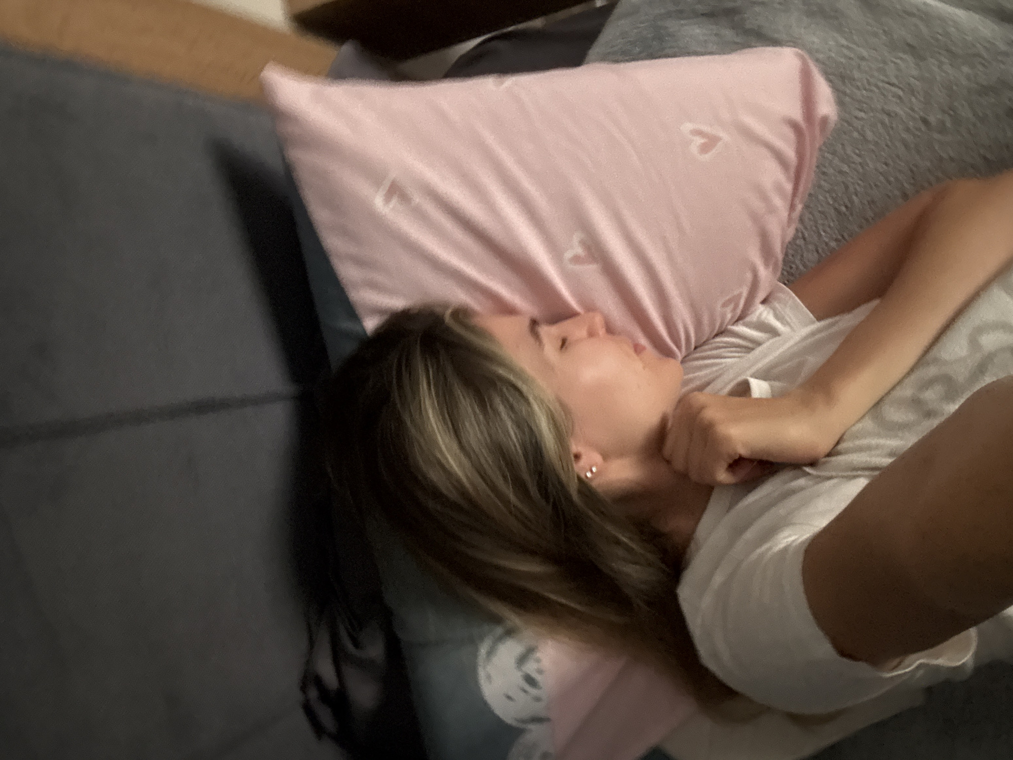 The image shows a young woman sleeping on a bed, with her head resting on a pink pillow adorned with small white hearts. She is wearing a white t-shirt. The bedding appears to be dark gray. The scene is dimly lit, suggesting it might be nighttime or indoors with low light. The woman is positioned on her side, with her right hand near her face. The background is slightly blurred but suggests a bedroom environment. No specific city or location details are visible in the image.