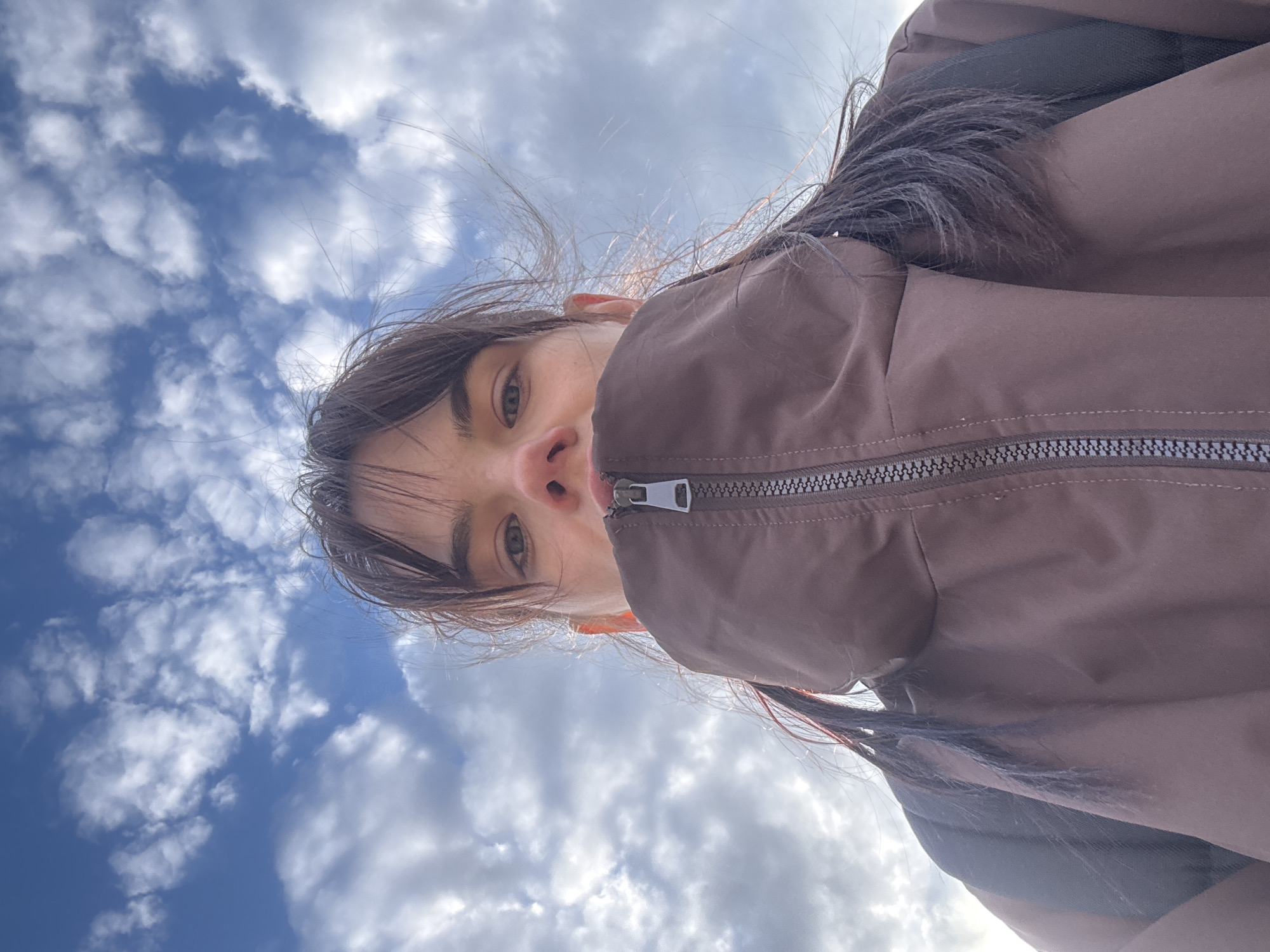 The image captures a young person from an extreme low-angle perspective, looking down directly into the camera against a bright, partly cloudy blue sky. The subject's face is positioned in the upper portion of the frame, with light-colored eyes (appearing greenish or hazel) and a neutral, direct gaze. Long, dark hair, some strands appearing windblown, frames the face and extends around the head. The person is wearing a brown or taupe-colored jacket with a prominent silver-grey zipper running down the front, visible on the right side of the frame. The jacket material appears smooth and possibly synthetic.
The background is dominated by a clear blue sky interspersed with numerous white, fluffy cumulus clouds, indicating fair weather during the daytime. The sun, while not directly visible, creates bright highlights on the clouds and edges of the hair. The overall composition suggests an upward-looking portrait or selfie, creating an immersive sense of being outdoors and looking towards the sky. The location context provided is Rostov-on-Don, Russia. There is no visible text in the image.