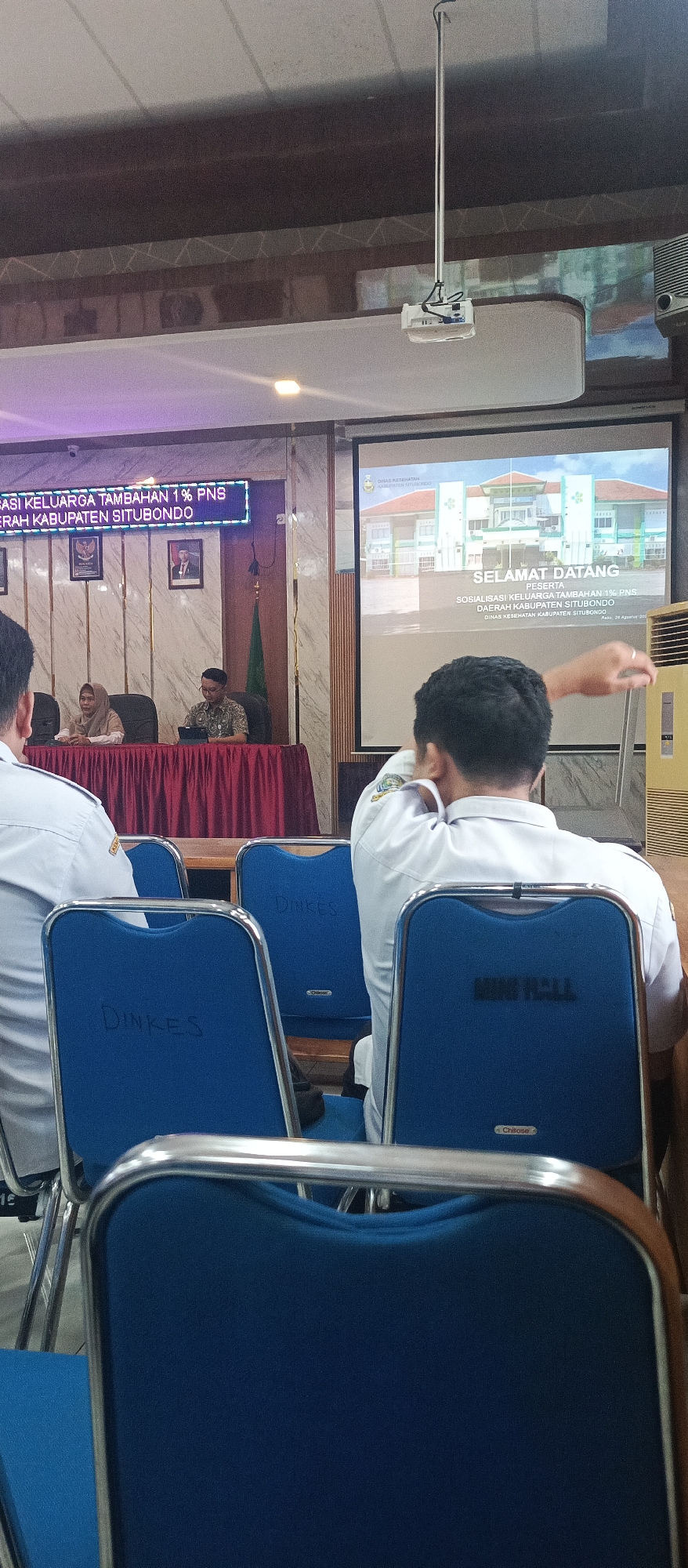 A group of people are seated in a room, attending a presentation. The room appears to be a meeting or conference hall, with a projector screen at the front displaying an image of a building and text. There are several rows of blue chairs, some with labels like "Dinkes" and "MING HALL." People are seen from behind, wearing white shirts. A man in a patterned shirt is seated at a table with a woman in a headscarf. The overall atmosphere suggests a formal event. The text on the screen and the signage on the chairs suggests the event is related to the health department ("Dinkes" likely being an abbreviation for "Dinas Kesehatan" or Health Department) and is taking place in Situbondo, Indonesia. The text on the projector screen reads "SELAMAT DATANG PESERTA SOSIALISASI KELUARGA TAMBAHAN 1% PNS DAERAH KABUPATEN SITUBONDO" which translates to "WELCOME PARTICIPANTS OF THE 1% PNS FAMILY SOCIALIZATION REGIONAL REGENCY OF SITUBONDO." Another line on the screen reads "DINAS KESEHATAN KABUPATEN SITUBONDO" which is "HEALTH DEPARTMENT REGENCY OF SITUBONDO." The date "Rabu, 26 Agustus 202" is also visible on the screen, indicating the event is taking place on Wednesday, August 26, 202_.