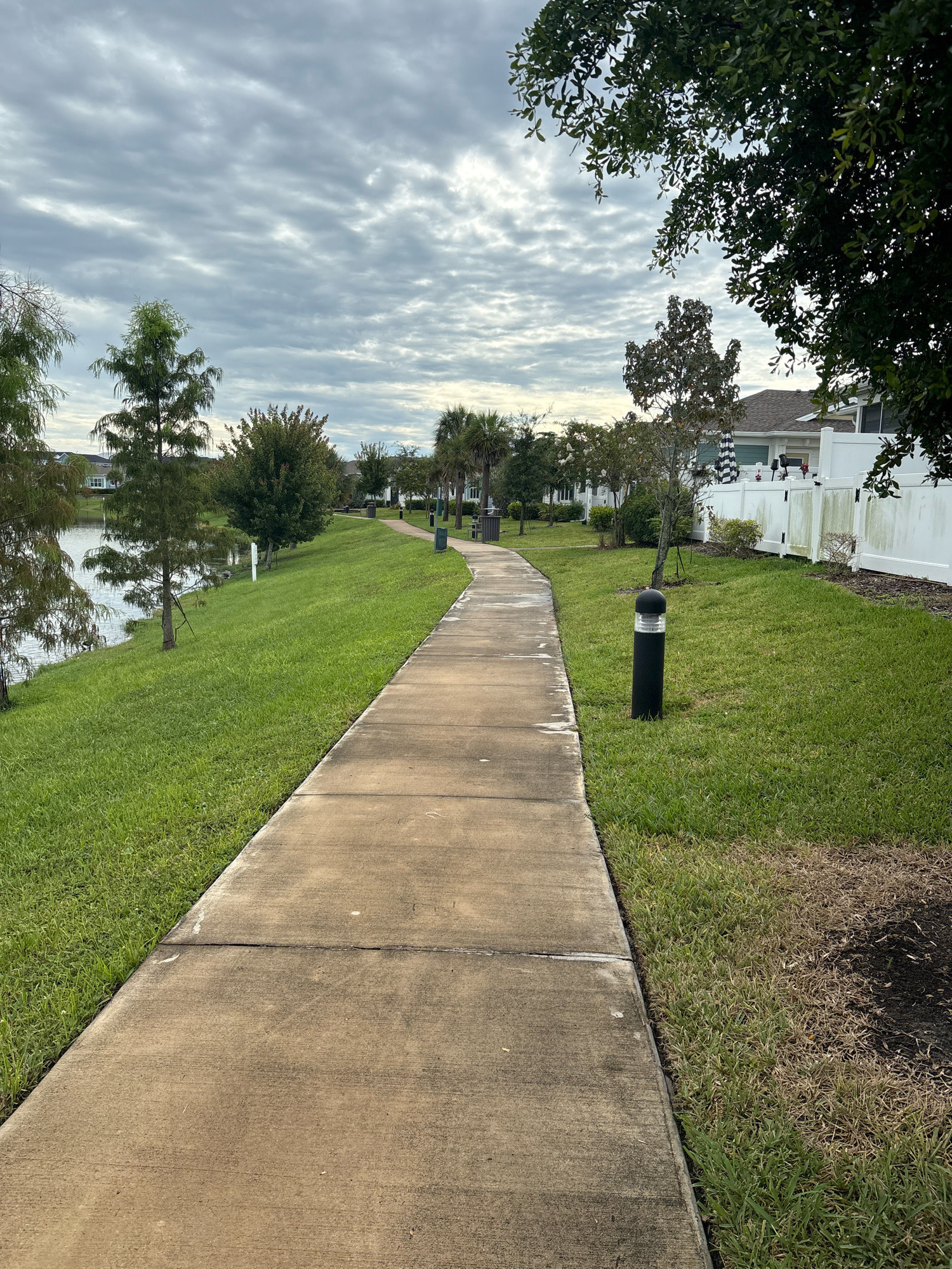 The image depicts a paved walkway curving alongside a grassy slope that leads down to a body of water on the left. The walkway is made of concrete, showing some signs of wear and staining. On the right side of the walkway, there is a well-maintained lawn with scattered black bollard lights. In the background, houses with white fences and mature trees are visible. The sky is overcast with dramatic cloud formations. The scene appears to be in a residential area with landscaping typical of a planned community or park. The specific location is not discernible from the image itself, but the environment suggests a warm climate.