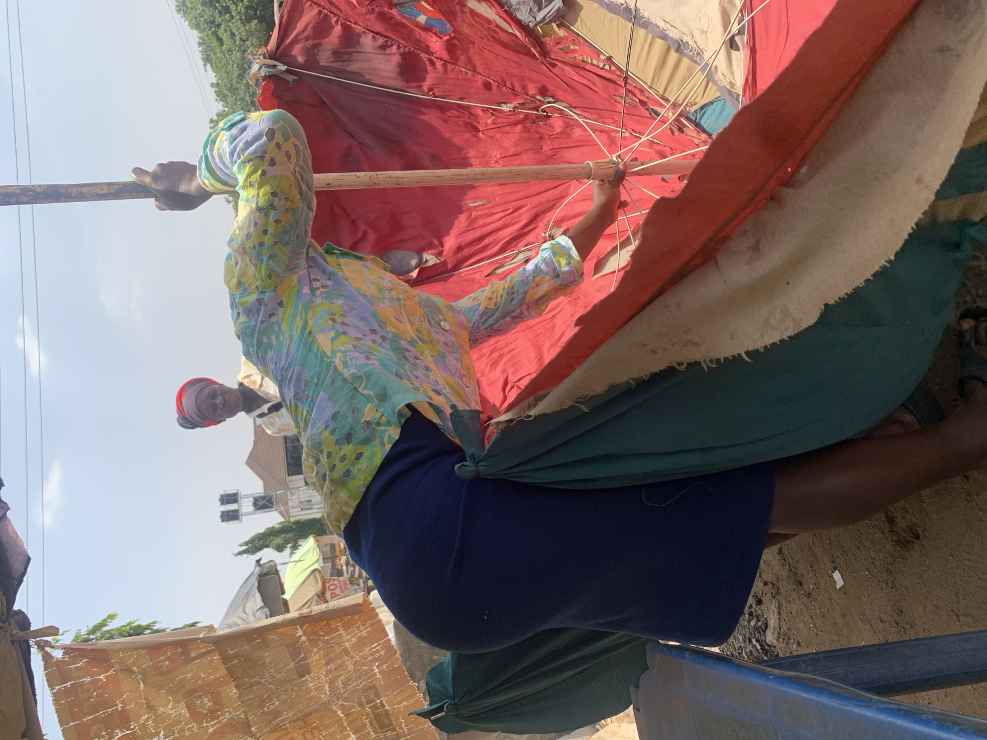In Abuja, Nigeria, a woman in a colorful patterned shirt and dark blue skirt is seen from behind, holding onto a wooden pole that appears to be part of a large, red umbrella. Her hand is wrapped around the pole, and she is actively engaged with the structure. Another woman with a red headscarf is visible in the background, looking towards the camera. The scene takes place outdoors, under a bright sky, with an overcast canopy overhead. The ground is sandy and uneven, suggesting a market or open-air space. Various makeshift stalls or structures, made of fabric and wood, are present in the background, along with some greenery and buildings. The overall impression is that of a busy, informal commercial area.