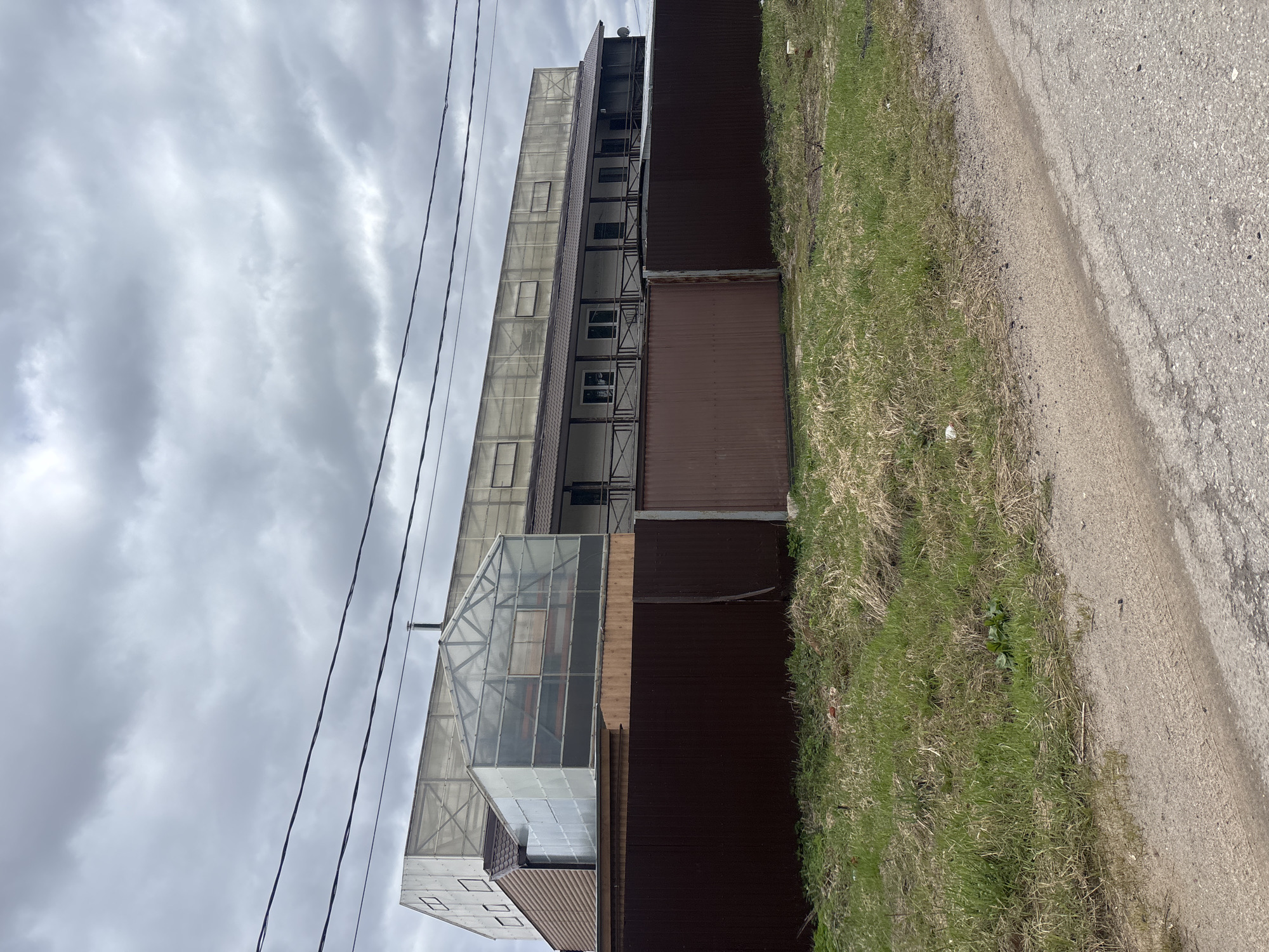 This image, taken in Раздолье, Russia, presents an exterior view of a functional building situated alongside an unpaved road, all under a heavily clouded sky. The photograph is oriented vertically, but depicts a landscape scene.
The central subject is a multi-story building characterized by a mix of materials. Its lower perimeter features a dark brown corrugated metal fence or wall. Above this, the main structure is clad in translucent panels, likely polycarbonate or similar material, supported by an exposed metal framework, giving it an industrial or large-scale greenhouse aesthetic. Multiple windows, some revealing internal elements and others reflecting the overcast sky, are interspersed across this facade. On the left side of the building, a prominent section constructed entirely of glass or translucent panels forms a distinct greenhouse-like addition.
Several thick, dark power lines stretch across the upper portion of the frame, positioned against the sky. To the right of the building's dark wall, a strip of mostly green grass mixed with dry, lighter vegetation separates the structure from a light gray, unpaved road or dirt path. The road surface appears worn, with visible cracks and loose gravel.
The sky is entirely overcast, filled with various shades of gray clouds, indicating a dull or non-sunny daytime. No people are visible, and no specific activity or event is captured, lending the scene a still and utilitarian character. There is no discernible text present within the image.