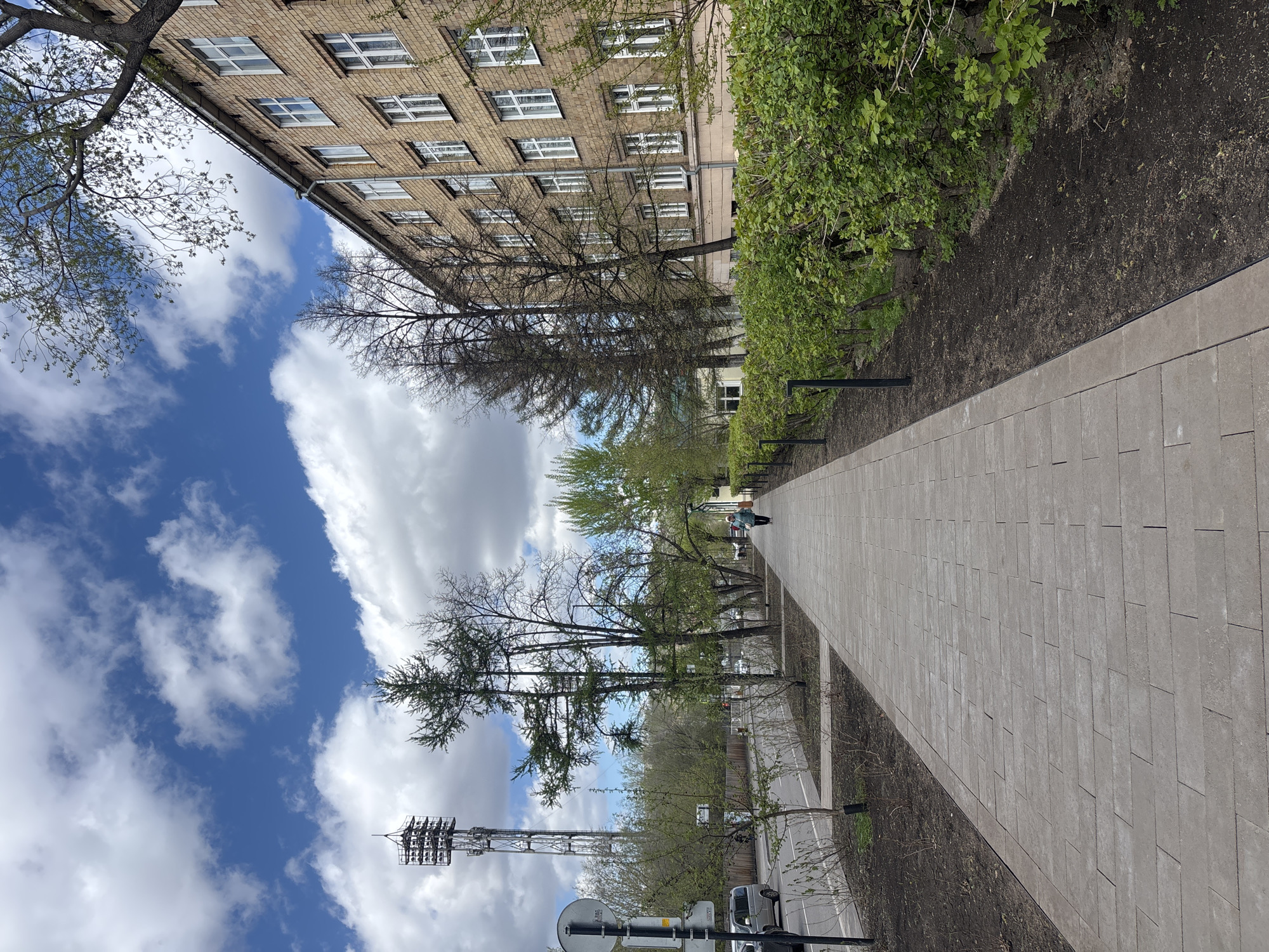 The image depicts an outdoor scene in Krasnoyarsk, Russia, featuring a paved walkway extending into the distance. To the left of the walkway is a road with vehicles, including a silver van, and a tall telecommunications tower. Above the road is a blue sky dotted with cumulus clouds. Adjacent to the walkway on the right is a narrow strip of uncultivated land bordered by dense green bushes and trees. A tan-colored brick building with multiple windows is visible along the top edge of the frame, partially obscured by trees. A person is walking along the walkway in the distance.