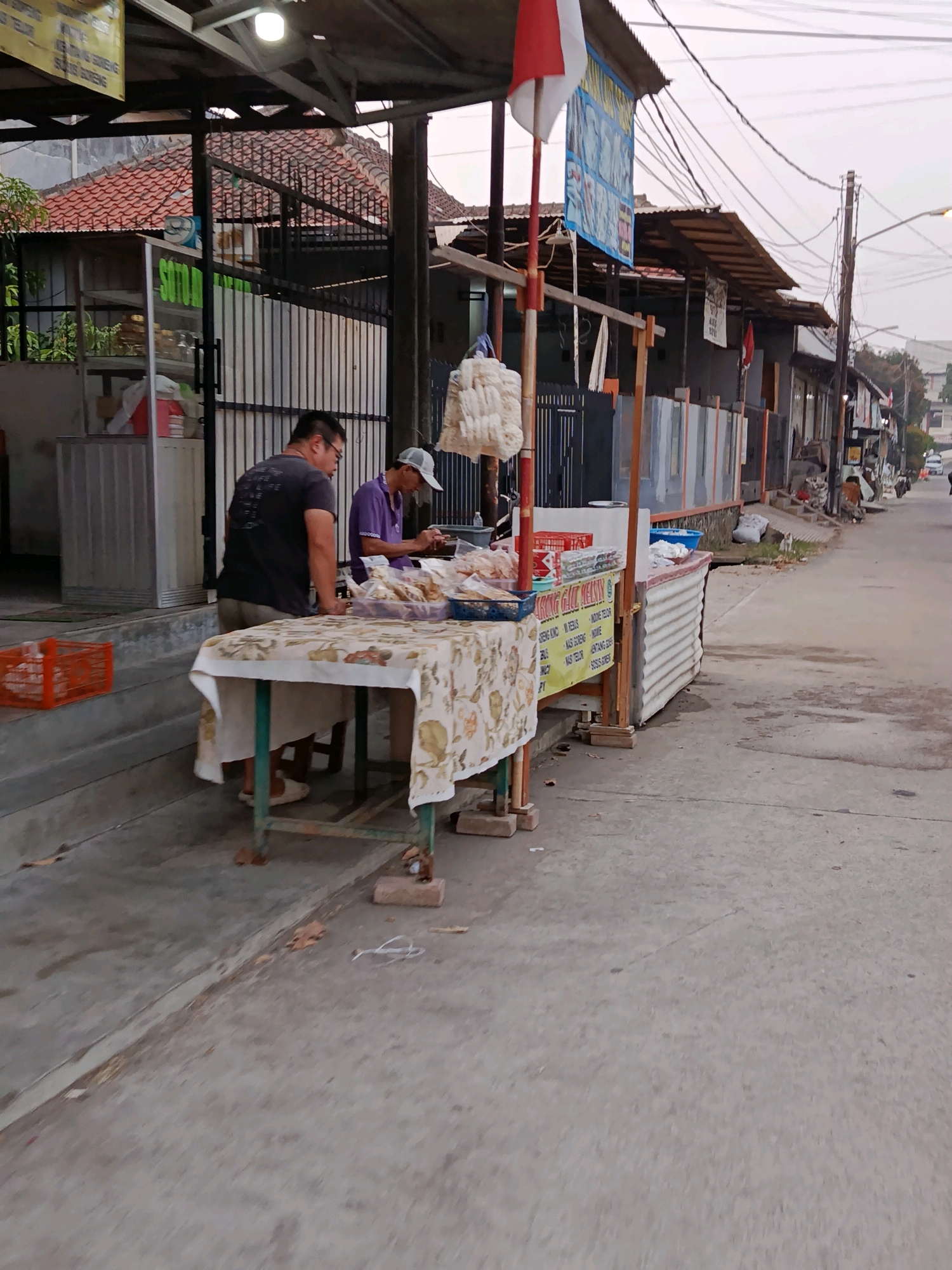 The image depicts a street-side food stall in Bekasi, Indonesia. Two individuals are visible at the stall, which is set up on a sidewalk next to a road. One person, wearing a dark t-shirt and glasses, stands facing the stall. The other person, in a purple shirt and a white cap, is behind the counter, appearing to be preparing or serving food. The stall features a table covered with a patterned cloth, displaying various food items in containers. Behind the stall, a building with a tiled roof and corrugated metal walls is visible, suggesting a commercial or residential area. The street is paved, and utility poles with numerous wires are present, common in Indonesian urban landscapes. The overall atmosphere suggests a local, everyday scene in Bekasi, likely during the late afternoon or early evening, given the lighting conditions.