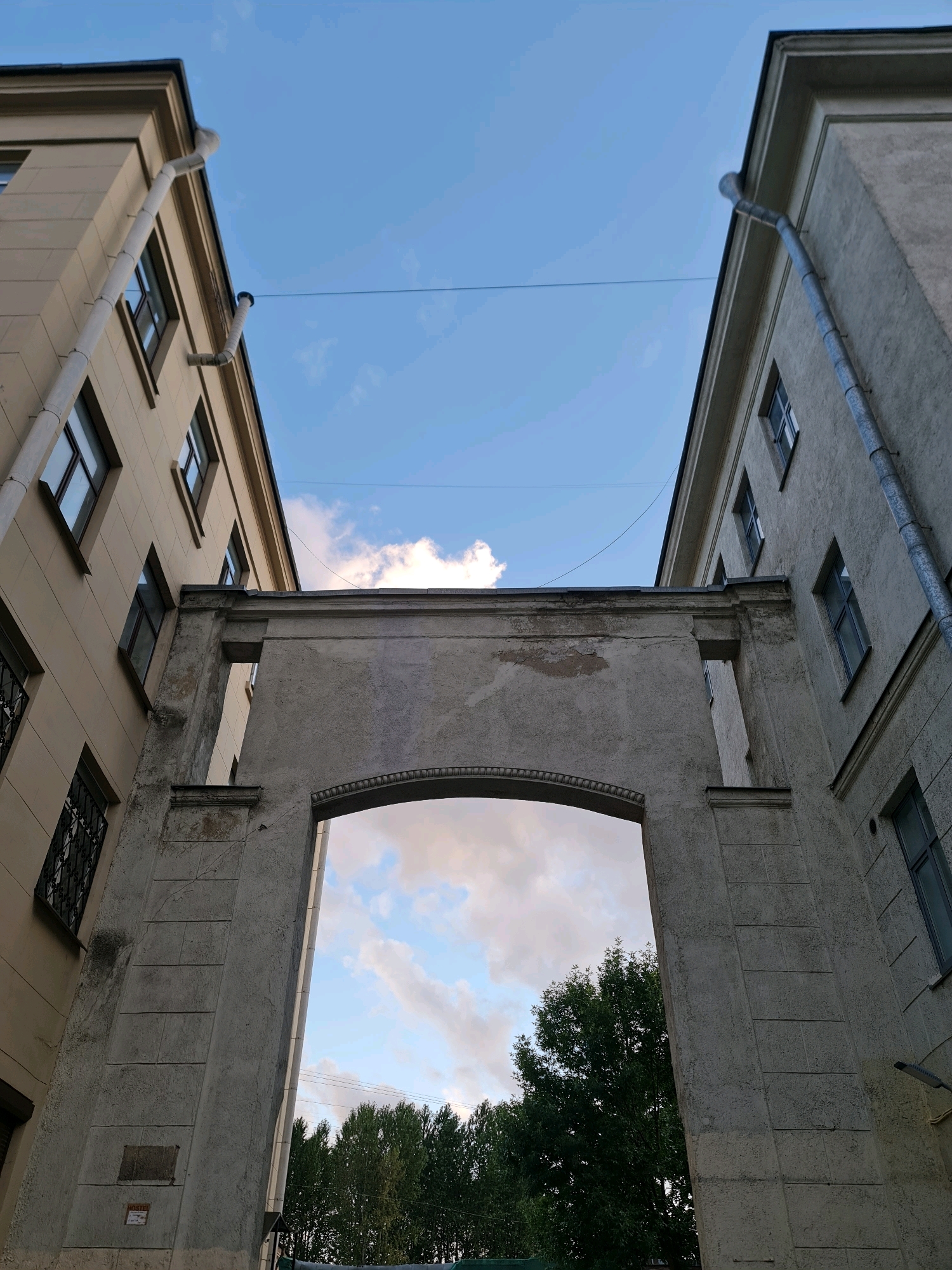 The image captures a low-angle view of an archway connecting two buildings in Saint Petersburg, Russia. The buildings appear to be old, with peeling paint on the archway and worn facades on the buildings themselves. The left building is painted a light beige, while the right building is a mottled gray. Both buildings have multiple windows with dark frames, some of which have protective metal bars. Gutter pipes are visible running down the sides of both structures.
Through the archway, a bright sky with scattered clouds is visible, along with the tops of lush green trees. A power line stretches horizontally across the frame, connecting the two buildings above the archway. The overall impression is one of an urban courtyard, likely in a historic district of the city, given the architectural style. The lighting suggests it is either early morning or late afternoon. No people are present in the image.