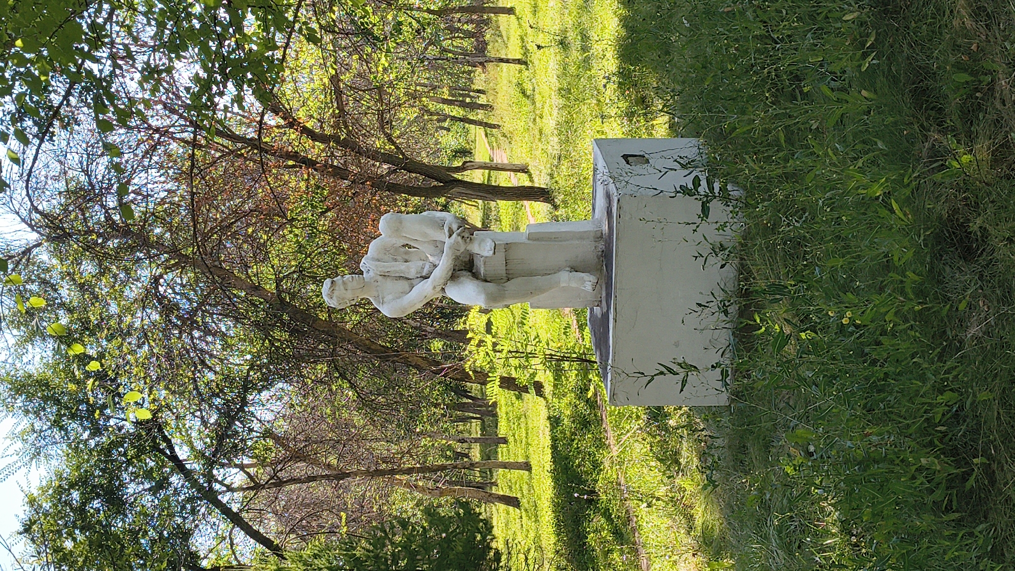 A white statue of a seated man holding a child is positioned on a pedestal in a park in Mikhaylovka, Russia. The statue is located in a grassy area with trees in the background. The man is depicted as muscular and is looking down at the child he is holding. The child is smaller and appears to be an infant or toddler. The statue is made of concrete or plaster and has a weathered appearance. The surrounding environment is lush and green, with sunlight filtering through the trees, creating dappled shadows on the ground. There is a pathway visible in the distance. The overall scene suggests a public park or garden, possibly a memorial or artistic installation.