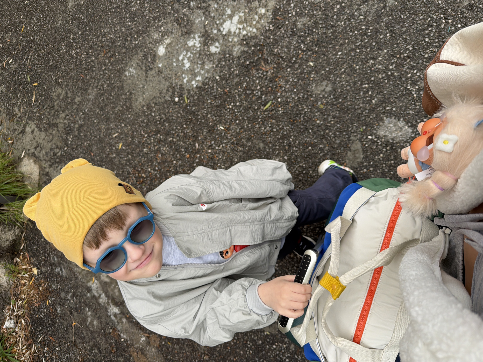 An overhead shot captures a young child looking up at the camera with a bright smile, outdoors in Nerin, Montenegro. The child, likely a toddler or preschooler, wears a vibrant yellow beanie with bear ears and stylish blue hexagonal sunglasses. They are dressed in a light gray jacket, dark blue pants, and athletic shoes (partially visible).
The child holds a silver and black remote control-like device in their right hand. Next to them, a light-colored backpack with a prominent orange zipper, blue, and green sections, and a yellow strap accent is visible. Two small plush toys are attached to the backpack or a nearby person: one resembles a baby doll, and the other is a fluffy animal adorned with a small white flower. A small, unreadable white tag with blue text is visible on the baby doll plush. Part of an adult's attire, including a brown and white hat and a white fluffy garment, is seen on the far right, suggesting an adult is accompanying the child.
The setting is an outdoor paved area, likely asphalt, speckled with white and a few scattered dry leaves. Patches of green grass and dirt are visible along the left edge. The diffused lighting indicates an overcast day. The child's happy expression and engaging gaze are notable.