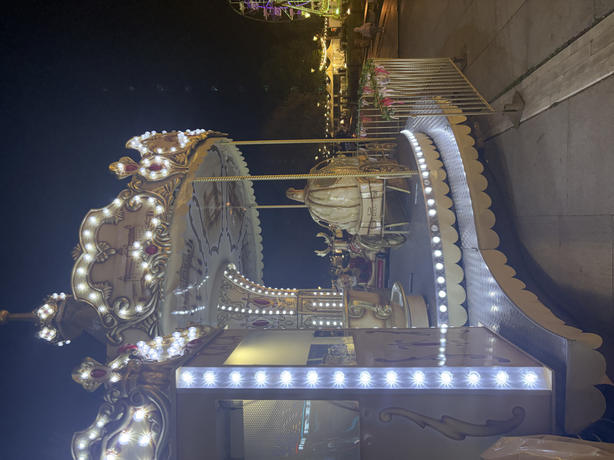 The image captures a brightly lit carousel at an amusement park or carnival at night. The main subject is the ornate carousel, which is lavishly decorated with white and gold accents and illuminated by numerous sparkling white LED lights, creating a festive and whimsical ambiance. Intricate scrollwork and possibly painted scenes adorn the carousel's upper sections. One prominent, highly detailed golden carriage-style seat is visible as part of the ride. The lower edges and base of the carousel are also lined with bright lights.
In the background, under a dark night sky, additional carnival attractions are faintly visible, including a Ferris wheel with purple and green lighting. Other distant lights suggest further rides or stalls. A few indistinct figures can be seen in the background, indicating the presence of people enjoying the event. The setting is clearly outdoors, with paved ground visible. The time of day is night, emphasized by the dark sky and the vibrant artificial illumination of the attractions. The scene conveys a lively and enchanting atmosphere typical of a fairground. There is no visible text in the image.