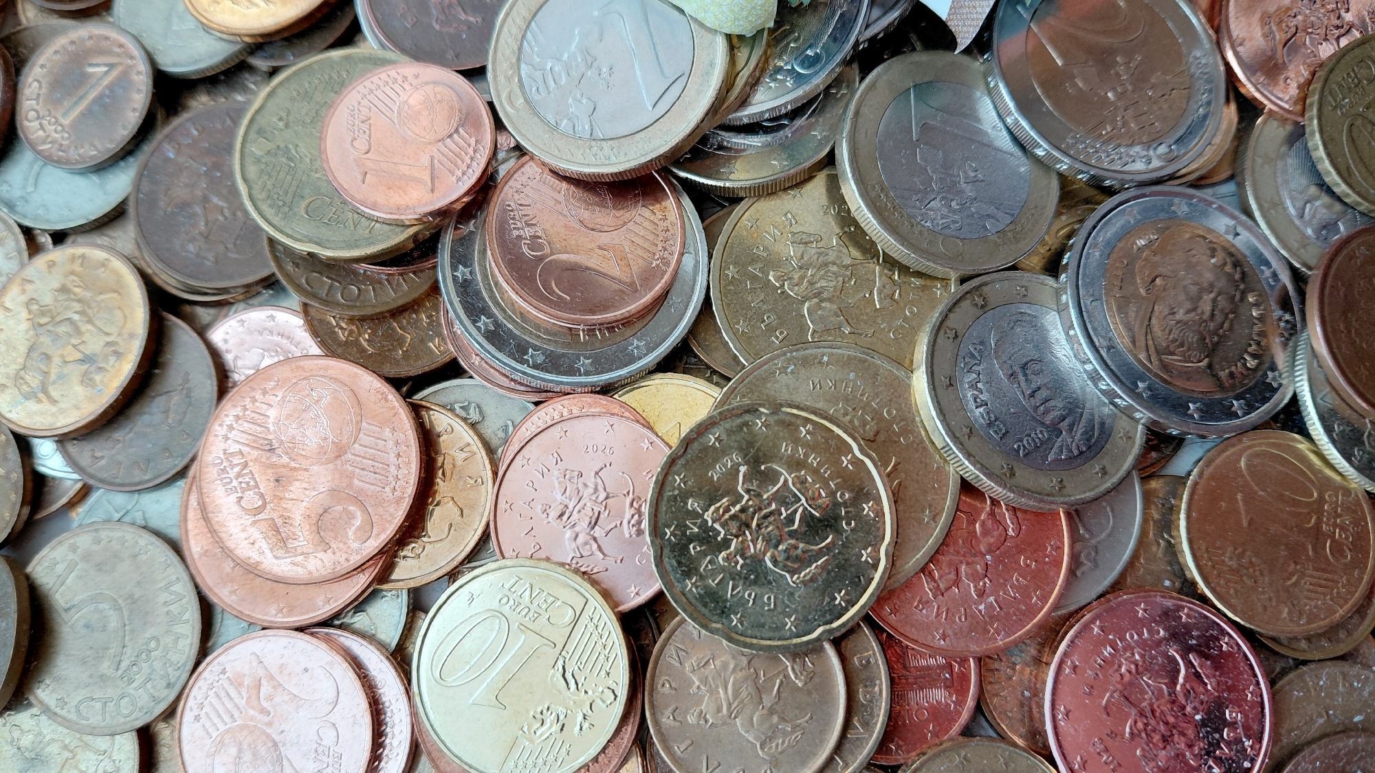 The image shows a close-up of a pile of Euro coins of various denominations. The coins are a mixture of copper, brass, and silver colors, with denominations including 1, 2, 5, and 10 Euro cents, as well as 1 and 2 Euro coins. Some coins have inscriptions in various languages, including Bulgarian, Spanish, and other European languages. The image is taken in a manner that the coins are closely packed together, making it difficult to distinguish the individual coins clearly. The location is unknown.