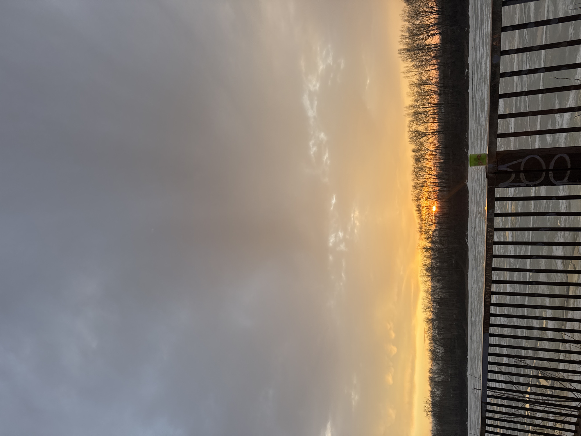 The image shows a sunset over the Kama River in Chaikovsky, Russia. The view is from a pier or walkway with a metal railing. The sky is a gradient of gray to orange as the sun sets behind a treeline on the opposite bank. A green cup sits on the pier next to the railing post with the painted number "200" is visible. The Kama River appears to be partially frozen or covered with ice.