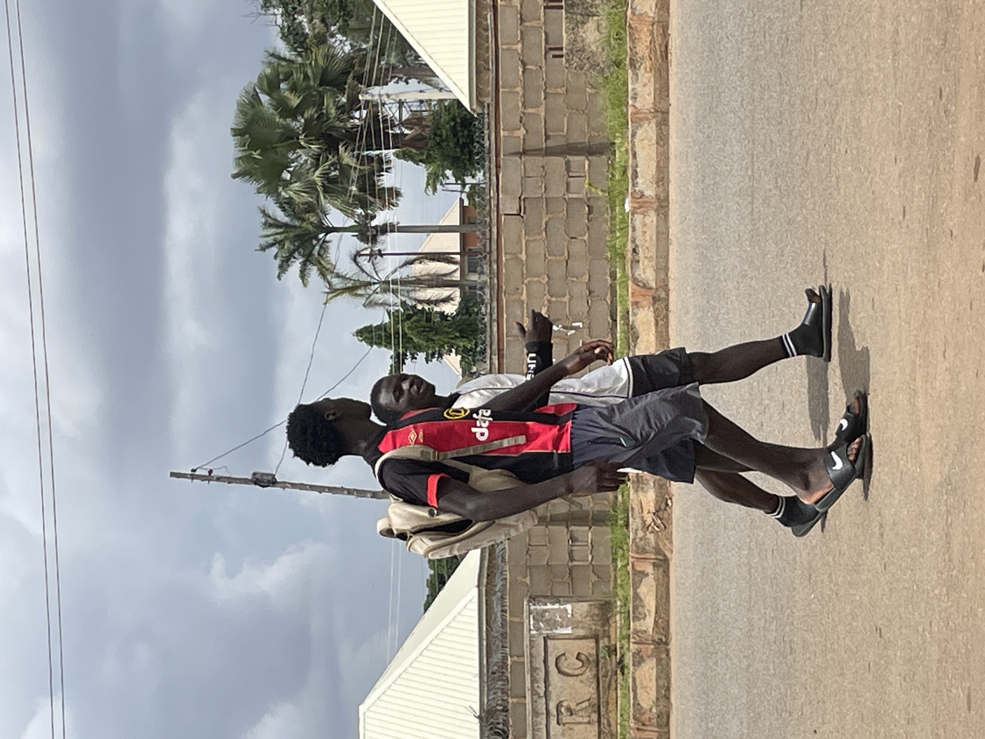 Two young Black men are walking side-by-side on a paved road in Bukuru, Nigeria. The man on the left wears a black and red soccer jersey with "dafa" written on it in white and a beige backpack. He has dark skin and short, curly dark hair. He is wearing dark grey athletic shorts and black slide sandals with white socks. The man on the right wears a white shirt and dark shorts. He has dark skin and short, dark hair. He is looking towards the man on the left and gesturing with his right hand. Behind them is a brick wall topped with barbed wire, and beyond that, several buildings with white roofs and lush green palm trees are visible. The sky is cloudy with a mix of gray and white. Power lines are strung across the sky. The ground is asphalt with some debris and a narrow strip of green grass along the edge of the road.