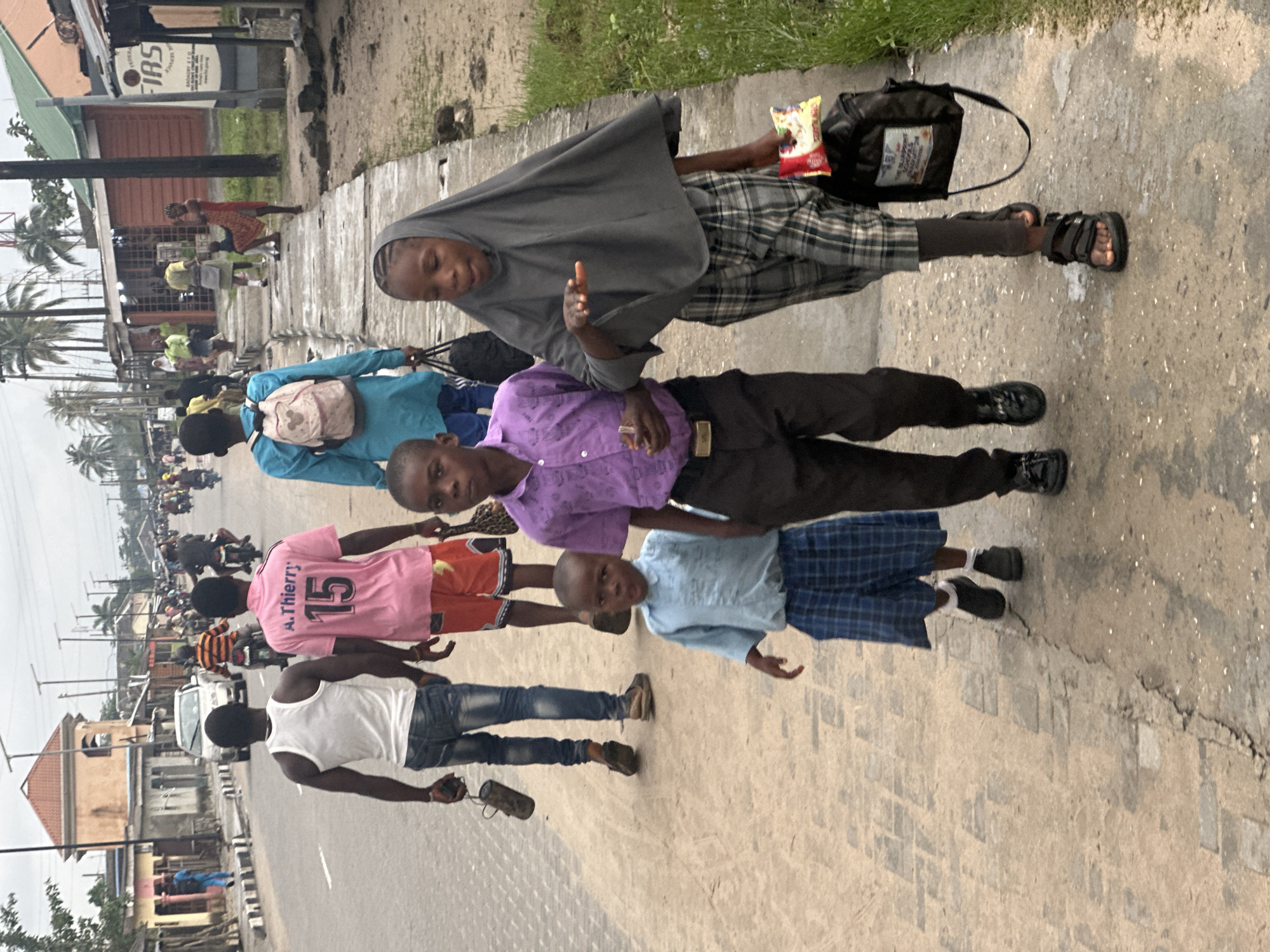 The image depicts a group of people, primarily children, walking along a sidewalk next to a street in Badagry, Nigeria. The scene appears to be outdoors in daylight, with overcast weather contributing to a diffused light.
In the foreground, a person wearing a grey headscarf and a plaid skirt walks ahead, holding a packet of snacks. They are followed by a child in a purple shirt and black pants, and another child in a light blue shirt and plaid shorts. Further down the sidewalk, a child in a pink t-shirt with the number "15" and "A. Thierry" printed on it walks with another child. A person in a white tank top and jeans walks behind them. The sidewalk is made of concrete and some brickwork, with a rough texture and visible cracks.
The street adjacent to the sidewalk is busy with traffic, including motorcycles and a white car. Buildings line the opposite side of the street, some appearing to be shops or houses. Palm trees are visible in the background, indicating a tropical or subtropical environment. The overall atmosphere seems to be one of daily life and movement in a bustling urban or semi-urban area. The people's expressions are not clearly discernible due to the image's orientation and resolution, but their presence suggests an activity like commuting, going to school, or simply moving through their community. The visible text on the pink shirt reads "A. Thierry 15". There is also text on a sign in the background, partially visible and appearing to be related to "FIRS".