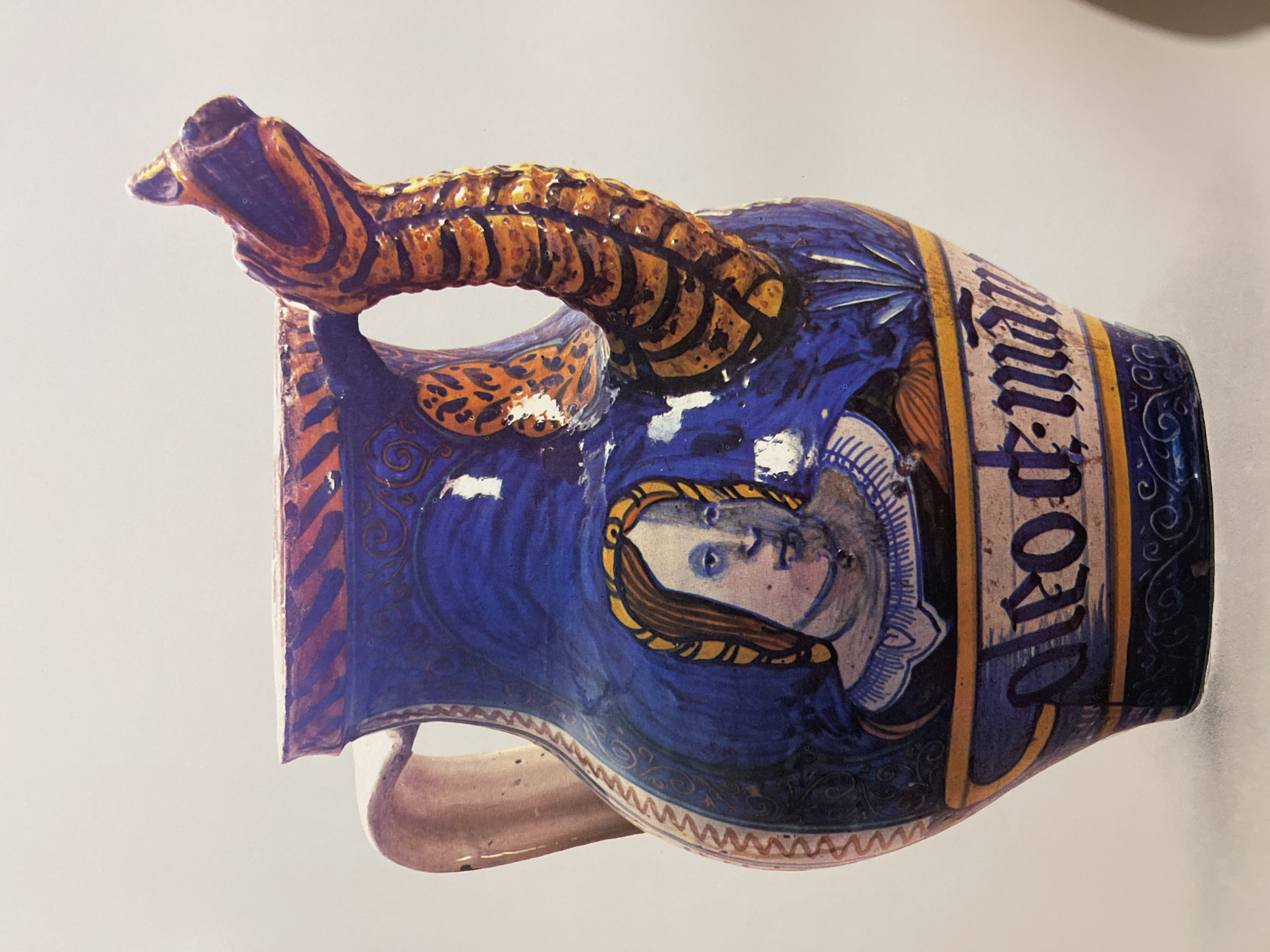 The image shows a ceramic jug. The jug is predominantly blue with a decorative handle shaped like an animal, possibly a dragon or lizard, colored in yellow and dark purple/brown. A portrait of a figure with pale skin and brown hair is painted on the side of the jug. There is a text inscription in a vertical format on the opposite side of the jug. The location is unknown.