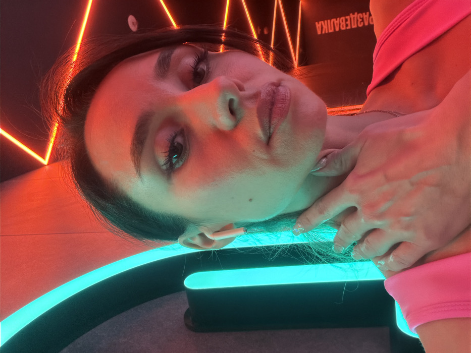 A close-up portrait depicts a female individual within a modern interior, identified as a changing room ("РАЗДЕВАЛКА" in Cyrillic, visible on the background wall), consistent with a location in Saint Petersburg, Russia. The individual has dark hair and dark eyes, wearing a bright pink top. A white wireless earbud is visible in her left ear. Her right hand is raised, with fingers gently touching her neck and jawline on the left side of her face. The scene is dramatically illuminated by contrasting light sources: a vertical, curved cyan-blue neon light on the left and angular red-orange neon light strips on the right, casting distinct colored light onto her face and the surrounding environment. The background walls are dark.