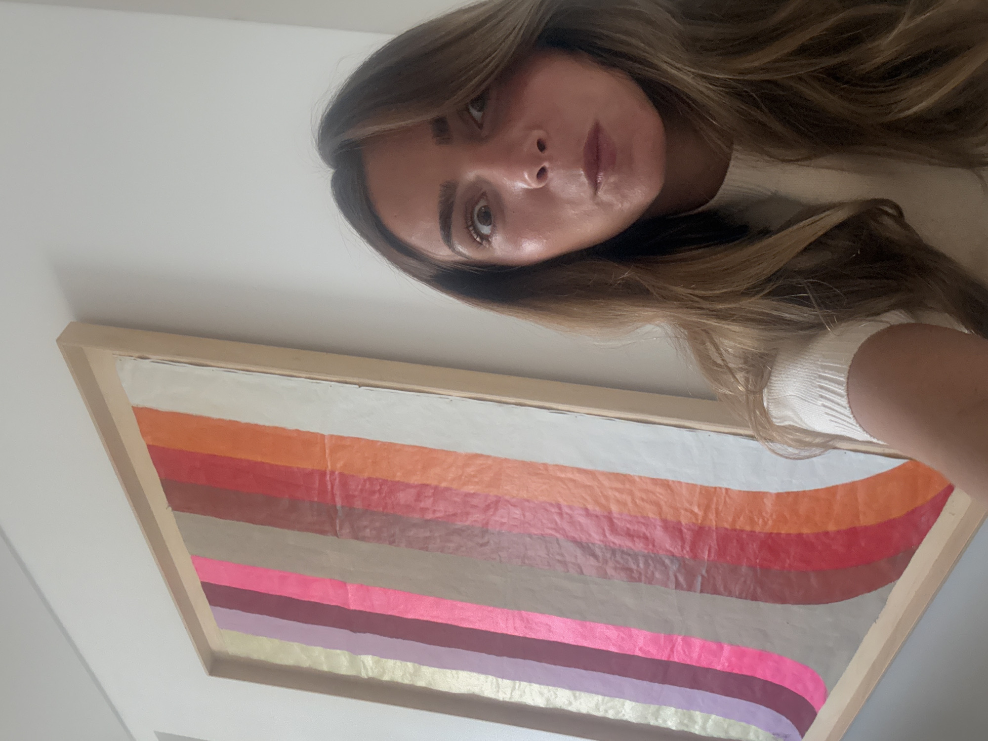 A woman is seen in a selfie-style photo, tilted towards the camera, with a piece of artwork visible in the background. The artwork features a series of horizontal, slightly curved stripes in varying colors, including orange, red, grey, pink, purple, and gold, within a wooden frame. The location is not determinable from the image.
