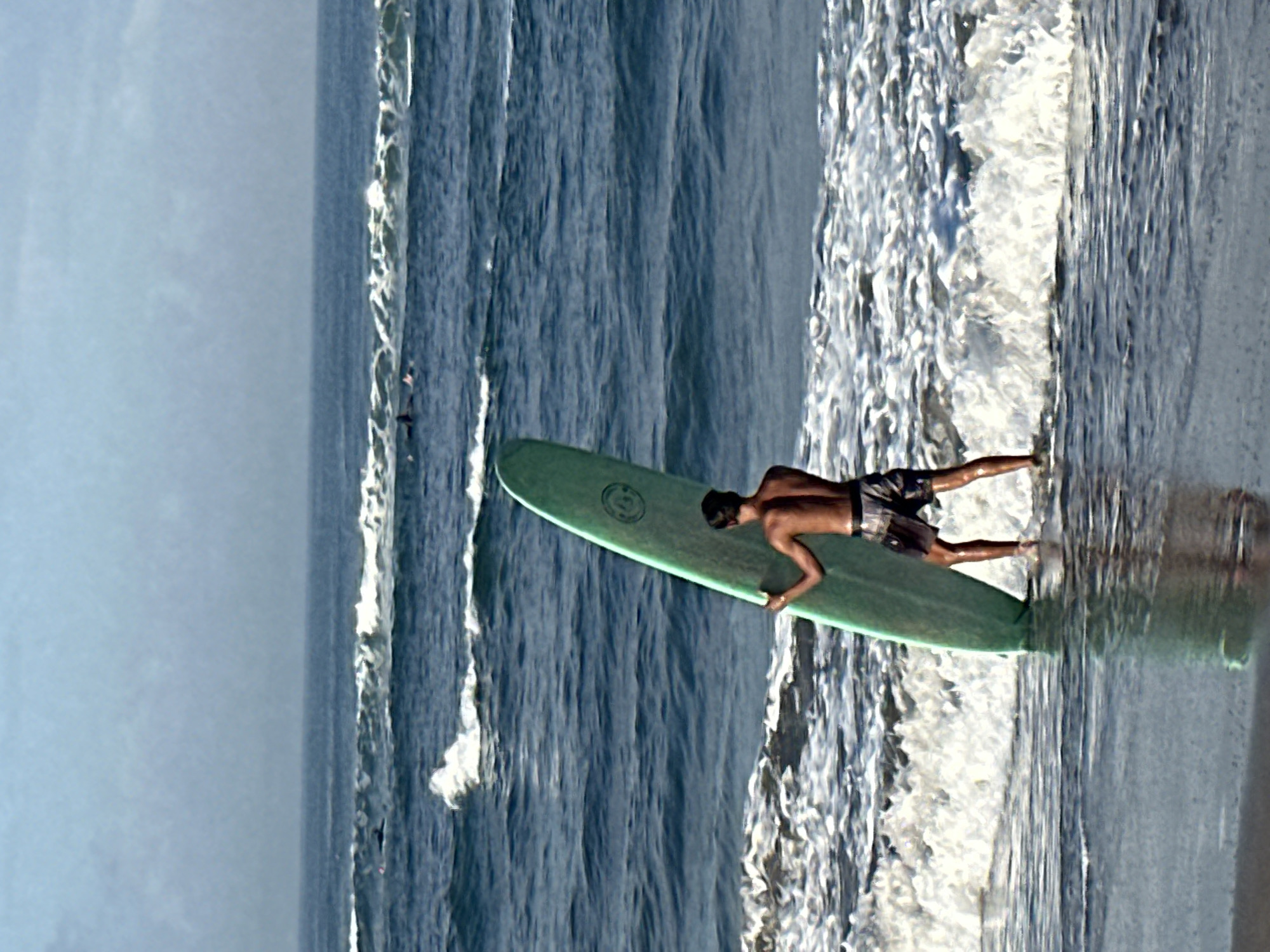A young man stands in the shallows of a beach, holding a light green surfboard upright. He is facing away from the camera, towards the ocean. The man is wearing dark patterned shorts. The ocean has moderate waves breaking on the shore. In the background, several other surfers can be seen in the water. The sky is overcast and hazy. The scene is likely at Batu Bolong Beach, a popular surf spot in Canggu, North Kuta, Indonesia.