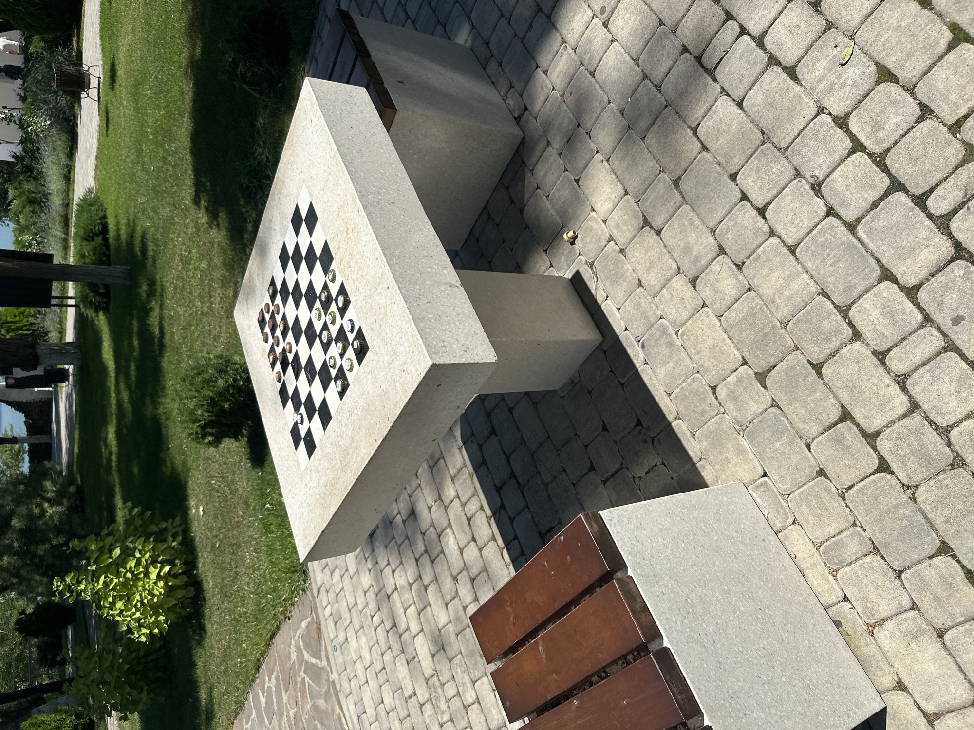 The image depicts a stone checkerboard table and bench in a park setting, likely in the city of Taman, Russia, given the context provided. The table, constructed from speckled grey stone, has a checkered surface on which checker pieces are arranged. To the left of the table, a bench with wooden slats is visible. The ground is paved with grey cobblestones. Beyond the paved area, a well-maintained lawn with scattered trees and bushes suggests a public recreational space. In the background, a body of water and distant structures are faintly discernible, hinting at a waterfront location. The scene is illuminated by natural sunlight, casting distinct shadows on the ground. No people are present in the image.