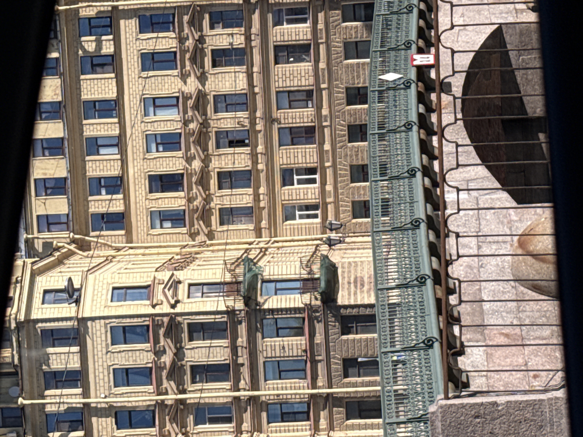 The image shows a building facade with multiple stories and numerous windows in Saint Petersburg, Russia. The building is made of light-colored brick and has decorative architectural elements. In the foreground, there is a bridge with a green ornate railing crossing a canal or river. Below the bridge, stone structures are visible, possibly part of the canal embankment or a tunnel entrance. The scene is captured from a slightly elevated perspective, with a dark frame or obstruction in the foreground and top of the image, suggesting the photo was taken from a vehicle or a window. The overall impression is of a historic urban landscape.
