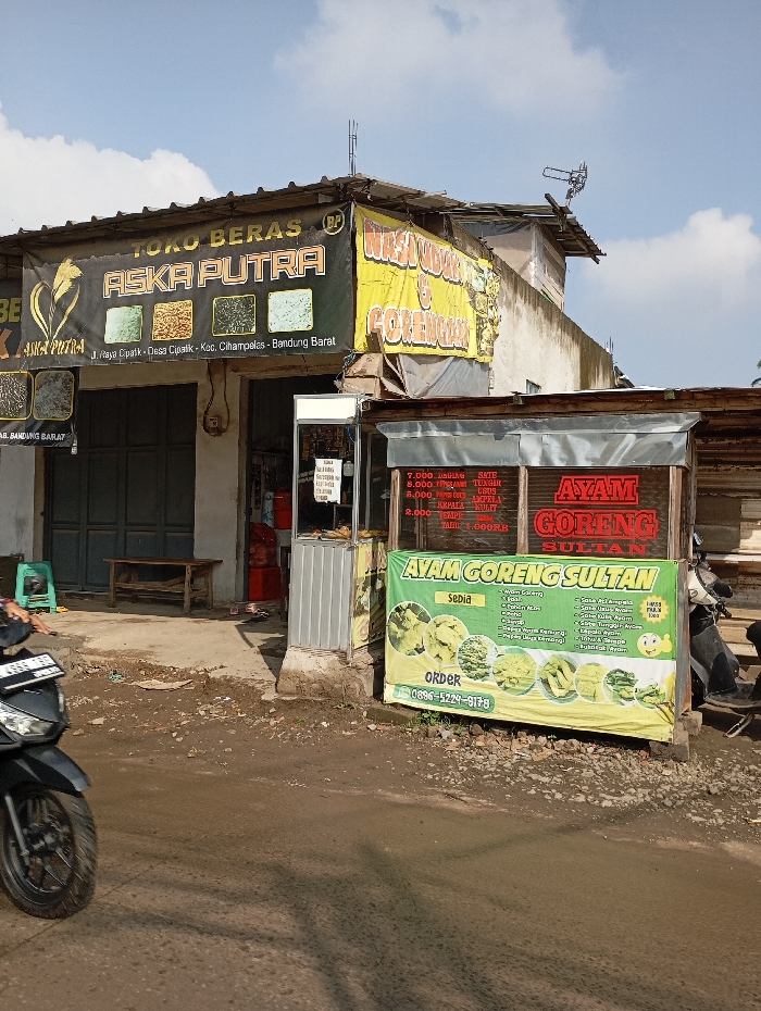The image depicts a static street scene in Situwangi, Cihampelas, Bandung Barat, Indonesia, under clear daylight conditions. The foreground features a rough, unpaved dirt and gravel surface. On the far left, the front wheel and part of the body of a black motorcycle with a partially visible Indonesian license plate "D 6866 TBP" are visible.
The midground is dominated by two adjacent commercial structures. To the left is "Toko Beras Aska Putra," a rice shop, housed in a building with dark, closed roll-up security doors. A wooden bench and a green plastic stool are positioned in front of these doors. Above the shop, a large black banner with gold text reads "TOKO BERAS ASKA PUTRA," displaying images of different rice varieties and the specific location "Jl. Raya Ciapatik - Desa Ciapatik - Kec. Cihampelas - Bandung Barat." A red gas cylinder is partially visible within the shop's entrance.
Immediately to the right of the rice shop is an "Ayam Goreng Sultan" (Sultan Fried Chicken) stall, a smaller, semi-permanent structure with glass display panels. Above this stall, a yellow banner partially visible behind the rice shop's banner reads "WARUNG NASI & GORENGAN" (Rice Stall & Fried Food). A prominent green and yellow banner is draped in front of the fried chicken stall, advertising "AYAM GORENG SULTAN" with illustrated menu items and an order contact number "0896-5229-9178." Prices for various food items are visible on the stall's glass, including "7.000," "8.000," "5.000," and "2.000." A building structure with an antenna mounted on its roof is visible behind the stalls. No individuals are present in the frame.