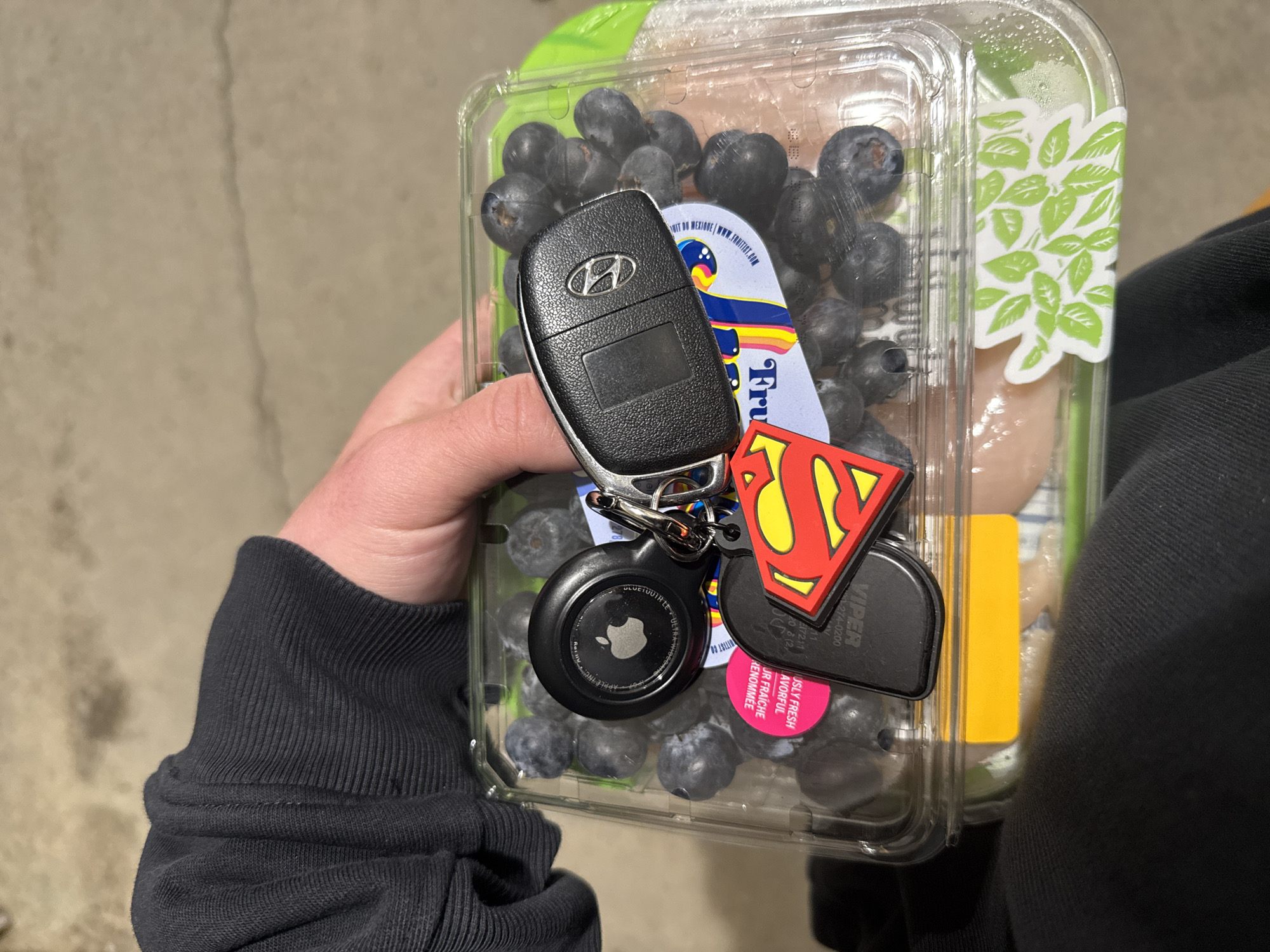 A person holds a clear plastic container of blueberries and white, round vegetables or fruits. A set of car keys hangs over the blueberries, featuring a Hyundai key fob, a black Apple AirTag, and a Superman logo keychain. The container has a green label with white leaves on the right side. The person wears a dark-colored sweatshirt. The setting appears to be outdoors on concrete. The location is not identifiable as Provincetown, United States of America from the visual details provided.