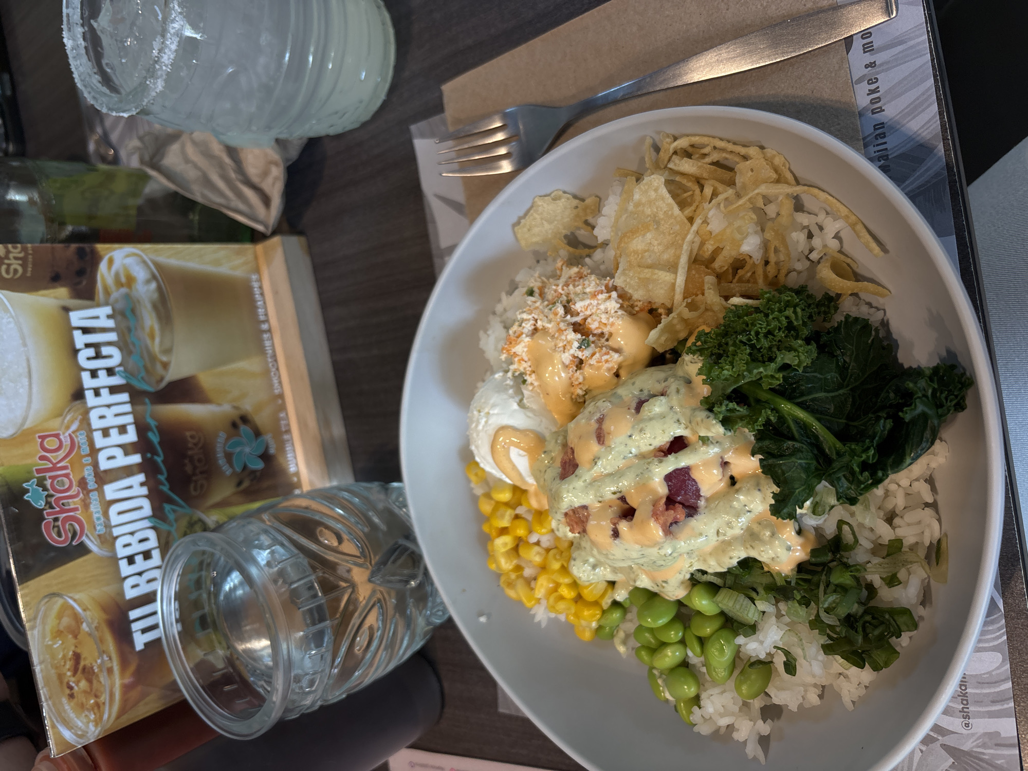 A close-up shot shows a partially consumed poke bowl on a table next to a glass with a salted rim, a glass with water, and a marketing booklet for "Shaka Hawaiian poke & more" in Mexico City, Mexico. The bowl features rice, corn, edamame, crab salad, fried toppings, kale, and two scoops topped with sauces. A fork rests on a placemat next to the bowl.