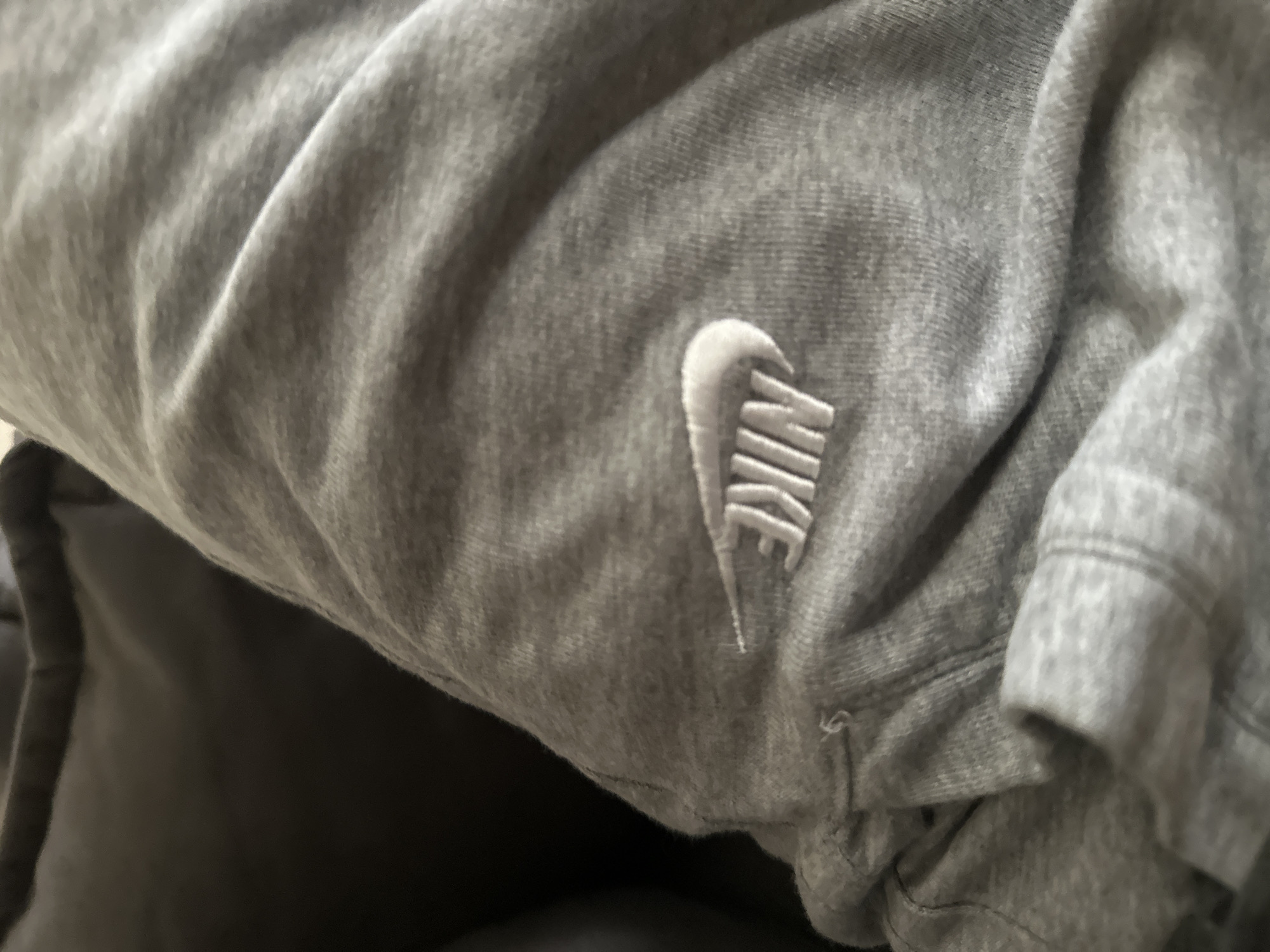 A close-up, high-angle shot shows a section of gray sweatpants with a white Nike logo embroidered on the fabric. The material is textured and appears slightly wrinkled, with a seam visible on the left side of the image where a small tear is present. The background is a dark, out-of-focus surface, possibly a couch or chair.