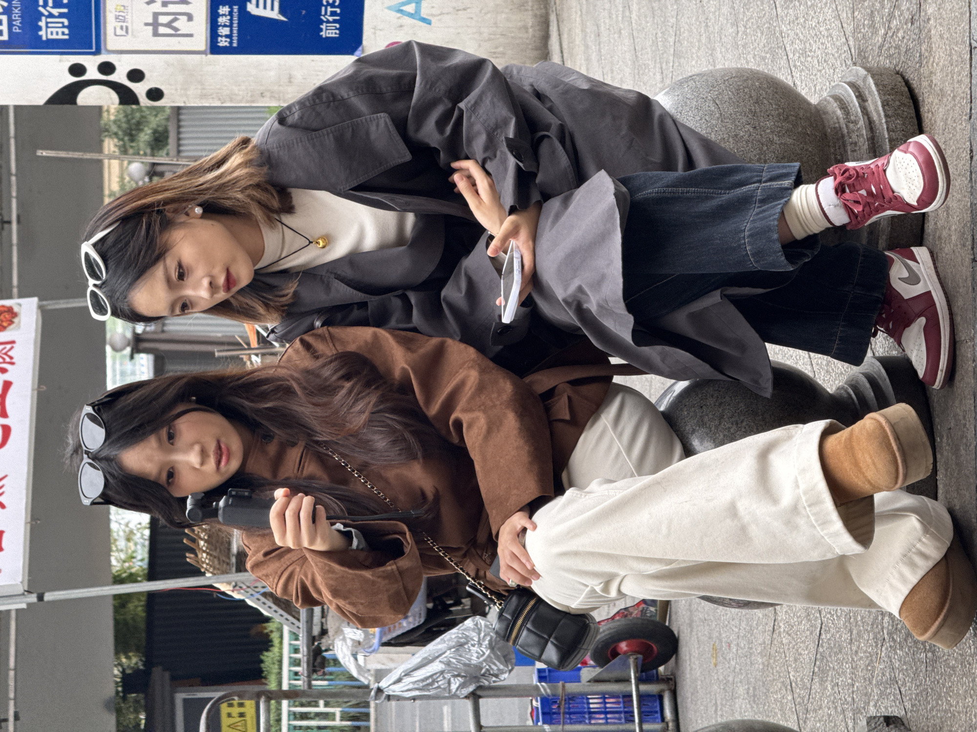Two young adult females are seated on cylindrical gray stone bollards on an urban sidewalk in Yuzhong District, China. The female on the left, with long dark hair and dark sunglasses perched on her head, is wearing a brown long-sleeved jacket, light-colored trousers, and light brown platform boots. She holds a black gimbal camera in her right hand, directing her gaze towards it. A small black quilted crossbody bag with a chain strap is visible on her right side. The female on the right, with medium-length ombre hair and white sunglasses perched on her head, is wearing a dark gray trench coat over a white top, dark blue jeans, and red and white sneakers with white socks. She holds a white smartphone in her left hand, and a gold spherical pendant hangs from her neck. Her gaze is directed towards the device held by the first female.
The background shows an urban environment with gray pavement. To the left, a metal railing, a yellow warning sign, and a partially obscured pushcart with blue crates and a silver tarp are visible. Directly behind the subjects, a light-colored banner with red Chinese characters hangs from an unseen structure. To the right, a blue and white street signpost displays Chinese characters, including "PARKING," "好省洗车" (Hao Sheng Car Wash), and directional arrows with "前行" (go straight/forward). Green foliage is discernible in the distant background.