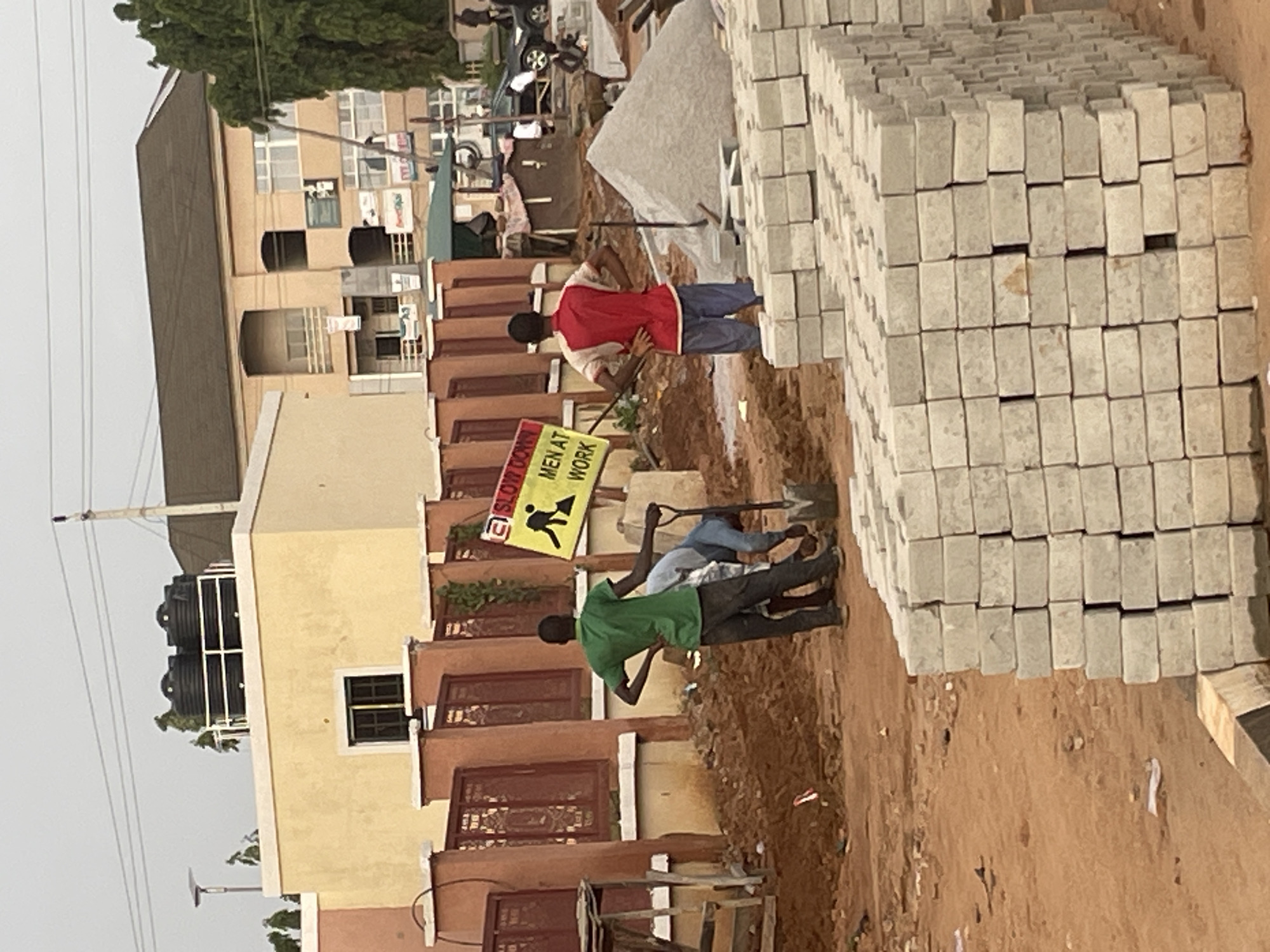 The image depicts a construction site in the city of Abuja, Nigeria. In the foreground, stacks of grey paving stones are arranged. Behind them, construction workers are engaged in labor on a dirt surface. One worker in a green shirt is standing with hands on hips, while another in a blue shirt is bent over, holding a shovel. A third individual in a red and white shirt stands observing the work. A yellow sign with the text "SLOW DOWN MEN AT WORK" is prominently displayed, featuring a silhouette of a worker with a shovel. The background reveals buildings, including a multi-story structure and a yellow building with water tanks on its roof, along with some trees and power lines, indicative of an urban setting.