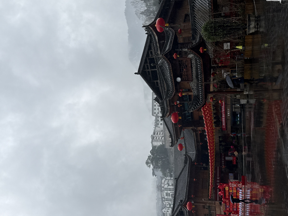 The image displays a traditional architectural complex in Gongtan, China, under an overcast sky. The ground, a paved square, is wet and highly reflective, suggesting recent or ongoing rain. Two individuals are visible in the foreground, each holding a black umbrella; one walks left-to-right on the left side, and another walks right-to-left on the right side. The buildings feature dark tiled, multi-tiered roofs with upswept eaves and appear to be constructed primarily of dark wood. Extensive red lantern decorations are present, including a long horizontal string of lanterns spanning across the square and individual lanterns hanging from various building eaves and balconies. A large red decorative display stands on the left side of the square. Signs with Chinese characters are visible on the buildings, including "古镇入口" (Ancient Town Entrance) above an archway on the right. A clock is affixed to the facade of the central building. In the background, modern multi-story buildings are situated on misty, tree-covered hills, with higher mountain ranges partially obscured by dense clouds.