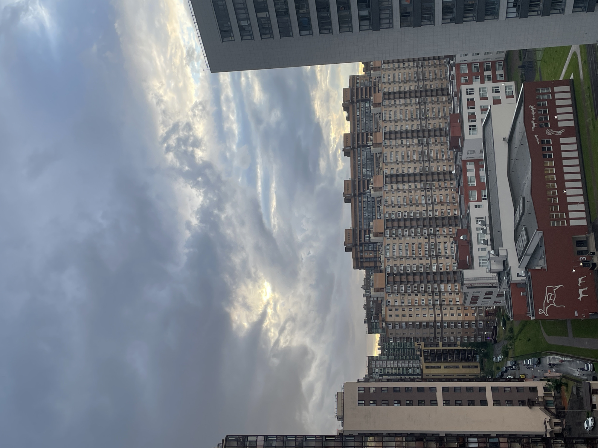 This is a vertical, eye-level, wide shot of a dense urban cityscape under a cloudy sky. The image is dominated by several tall residential buildings of varying architectural styles and colors, closely packed together. On the left, a pale grey, modern building with many rectangular windows stands prominently. To its right, a series of older, taller buildings with light brown and beige facades, featuring numerous balconies and windows, stretch into the distance. Further to the right, a prominent red-brown building with white framed windows and a distinctive white line drawing of what appears to be a cow or a similar animal is visible. Between and around these buildings, green grassy areas and a winding road with parked cars are visible, suggesting a residential neighborhood. The sky is filled with dramatic clouds, with patches of sunlight breaking through, indicating either sunrise or sunset. The overall mood is somewhat overcast but with hints of warmth from the sun. There is no visible text in the image.