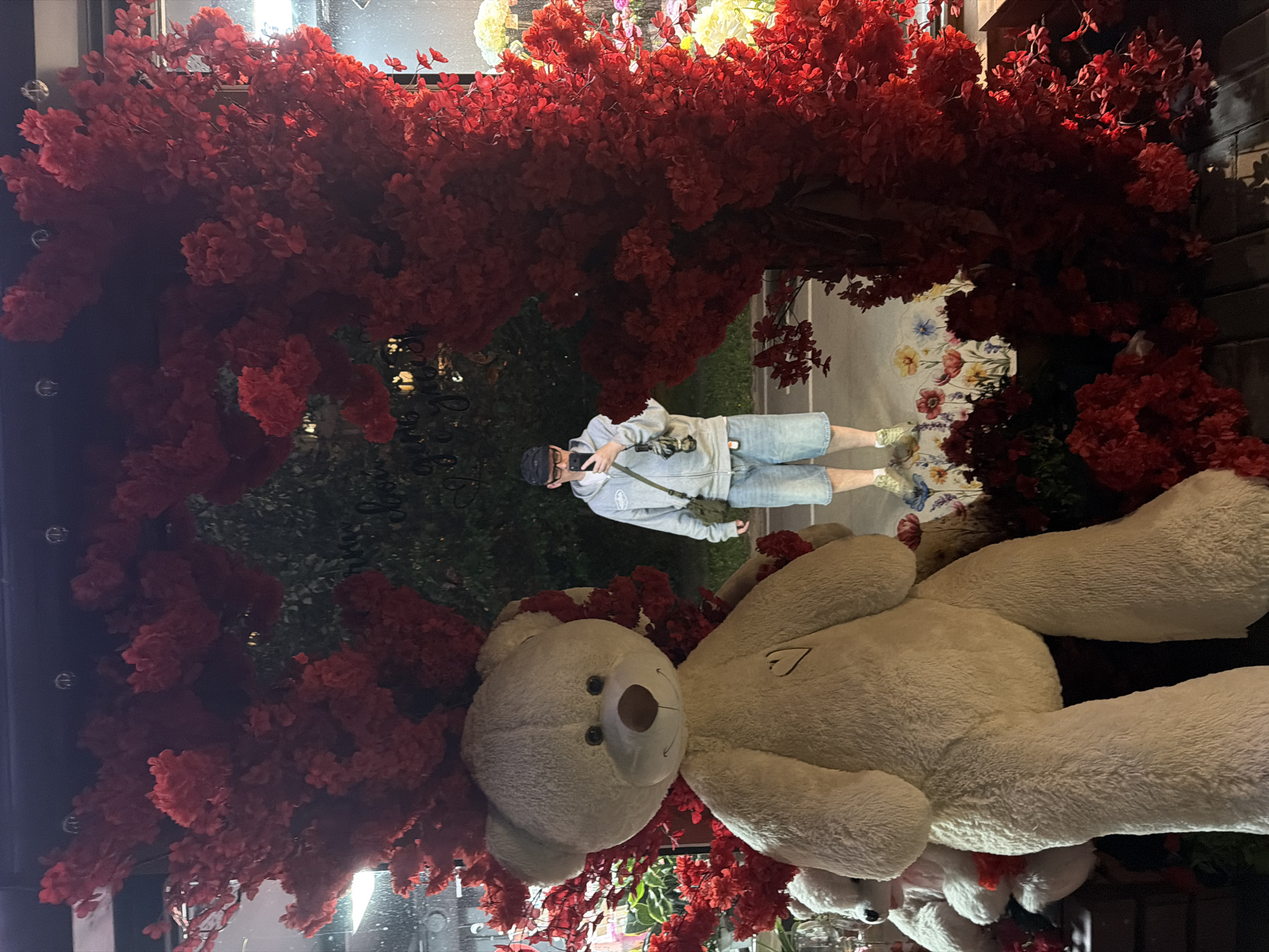 In Khimki, Russia, a person, wearing a black cap, glasses, a grey zip-up hoodie, and denim shorts, is standing in front of a large display. The display features an oversized cream-colored teddy bear positioned on the left side of the frame. Behind the teddy bear and to the right, the person is reflected in a mirror. The mirror is framed by a dense arrangement of red artificial flowers, creating a dramatic backdrop. The person is holding a mobile phone and taking a selfie. To the left of the main teddy bear, a smaller, white teddy bear is partially visible, also adorned with red flowers. The ground in front of the display is covered with a floral-patterned rug. The overall setting appears to be a shop window or a decorative installation.