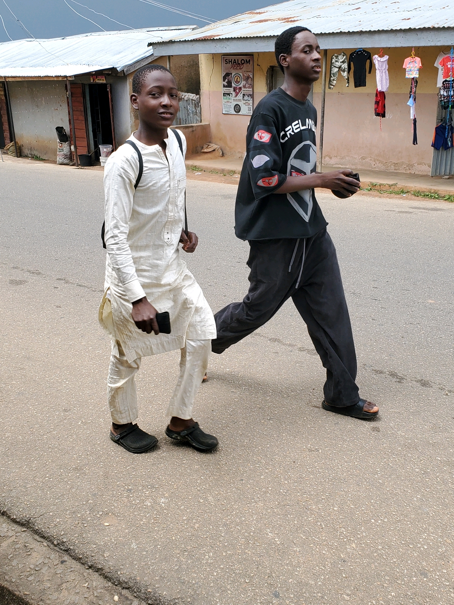 Two young males are walking on a paved road in Jos, Nigeria. The male on the left is wearing a white traditional tunic and trousers, and black clogs. He is carrying a black bag over his shoulder and holding a black phone in his right hand. The male on the right is wearing a black t-shirt with a graphic design and dark gray sweatpants, along with black sandals. He is holding a dark object, possibly a wallet or phone, in his right hand. In the background, there is a building with a corrugated metal roof, storefronts displaying clothing, and posters on the wall. The road surface is asphalt with some visible cracks and debris. The sky is overcast.