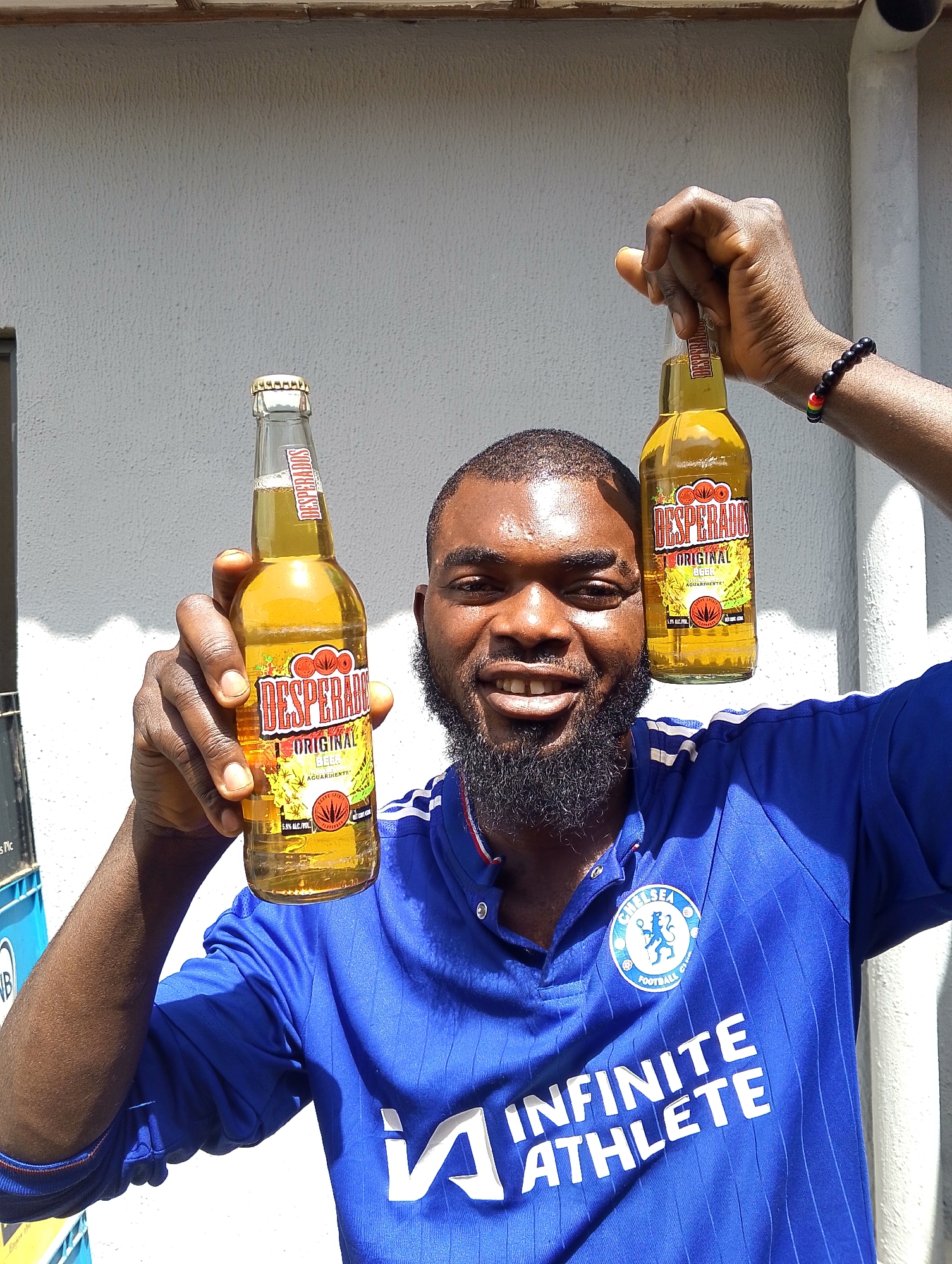 This is a medium shot, outdoor photograph taken in Jos, Nigeria. A Black man, smiling broadly, is holding up two bottles of Desperados beer. He is wearing a blue Chelsea Football Club jersey with white stripes and the club crest, and the words "INFINITE ATHLETE" visible on the front. The man has a short, dark beard and short black hair. One bottle is held in his right hand, and the other in his left. The bottles are mostly full of a golden-colored liquid, and the labels are clearly visible, reading "DESPERADOS ORIGINAL BEER AGUARDIENTE" and indicating 5.9% alcohol by volume. He is wearing a beaded bracelet with red, yellow, and green colors on his right wrist.
The background consists of a textured white wall and a thin wooden ledge at the top. The lighting suggests it is daytime, with bright sunlight casting shadows. The man's expression is jovial and celebratory. The scene appears to be casual and social. Visible text on the bottles and jersey includes brand names and logos.