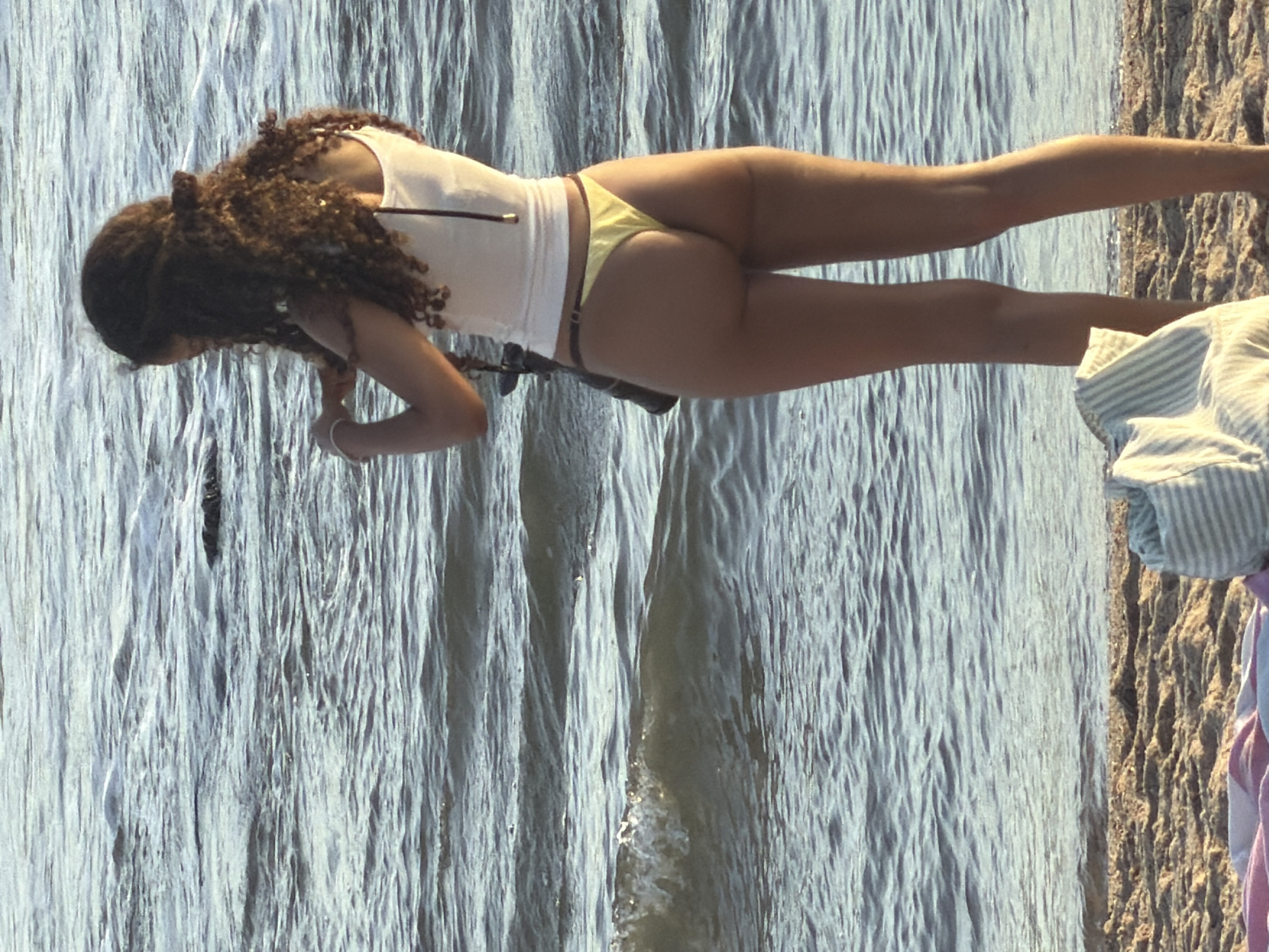 A young woman with curly dark hair stands on a sandy beach facing the ocean, wearing a white tank top and yellow bikini bottoms. She is holding a camera and appears to be taking pictures. The ocean has gentle waves. A towel and bag are visible on the sand in the foreground. The scene suggests a sunny day at the beach. The location is likely the coast of Espinho, Portugal, known for its beaches and surf.