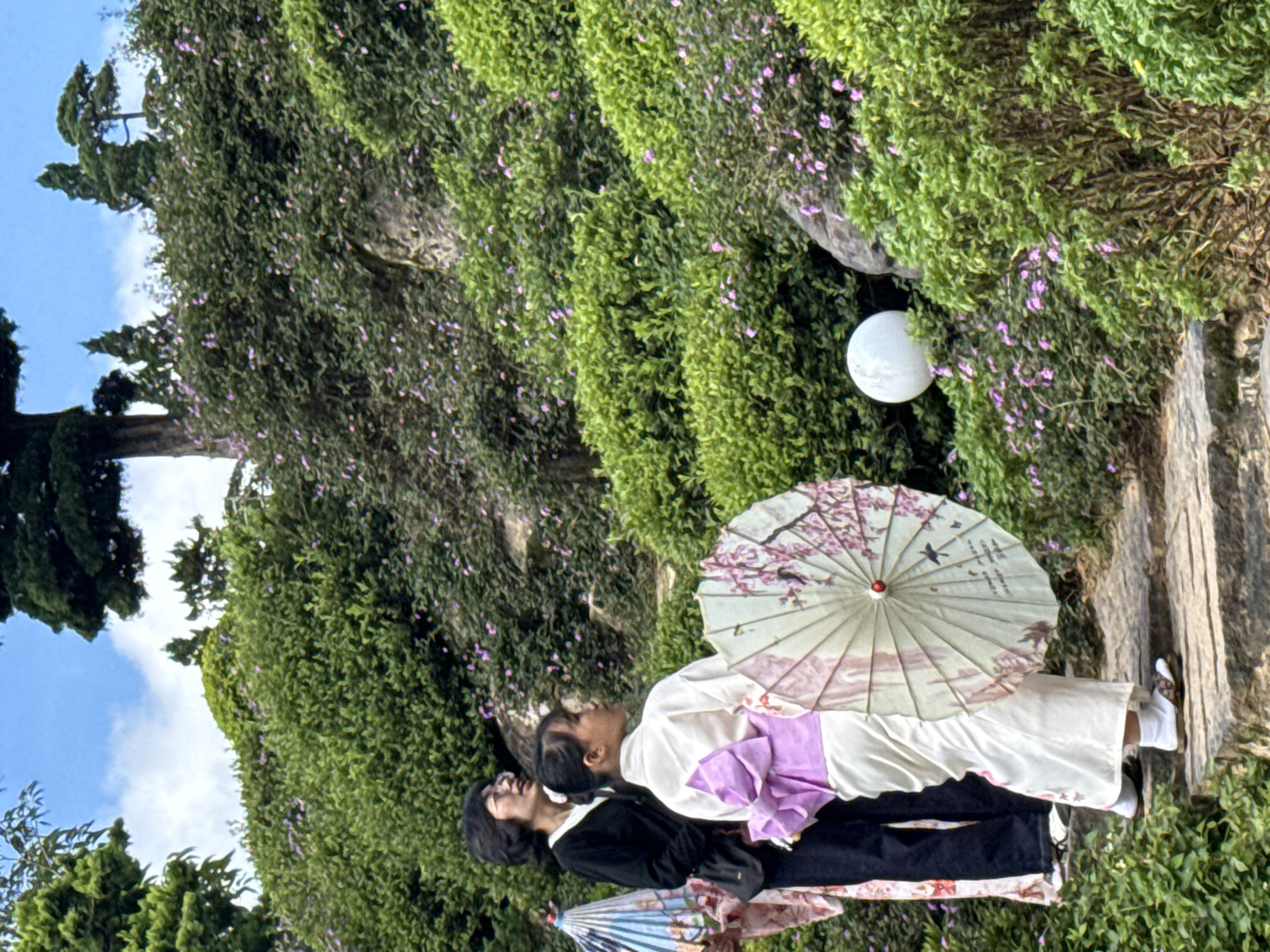 The image displays a garden scene in Cam Ly - Đà Lạt, Vietnam, featuring two women on a stone-stepped path surrounded by abundant greenery. In the foreground, a young woman is positioned with her back partially turned, wearing a white kimono or yukata secured with a prominent lavender-colored obi tied in a large bow at her lower back. She wears white tabi socks and open-toed sandals. She holds a large, round, light-toned parasol, embellished with cherry blossom patterns and a bird design, tilted slightly away from the viewer. Her gaze is directed upwards and to her right.
Standing behind and slightly to the left of the first woman is another woman with dark, short hair and glasses. She wears a black cardigan over a light-colored top and dark trousers. Her head is tilted upwards, and she is looking in the same general direction as the woman in the kimono. Partially visible on the far left edge of the frame, another individual's lower body in a patterned garment is seen, along with a blue and white patterned parasol.
The background is dominated by a lush, green hillside densely covered with various shrubs and bushes, dotted with numerous small purple flowers. Several larger trees, some with thick dark trunks and others resembling conifers, rise above the foliage against a bright blue sky with scattered white clouds. A prominent spherical white garden light is situated among the bushes on the right side of the stone path. The path itself consists of irregular flat stone steps.