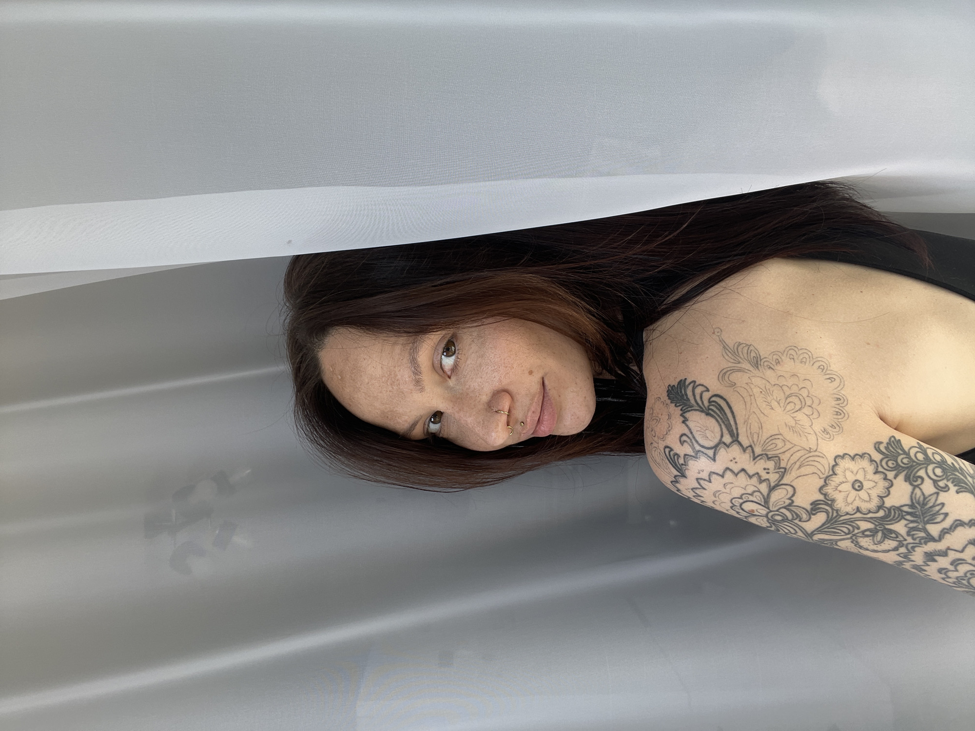 A woman with auburn hair, freckles, and multiple piercings in her nose is lying on a surface covered with translucent white fabric. Her left arm is visible and features a floral tattoo extending from her shoulder to her forearm. The city of Якшур, Russia is not discernible from the image.