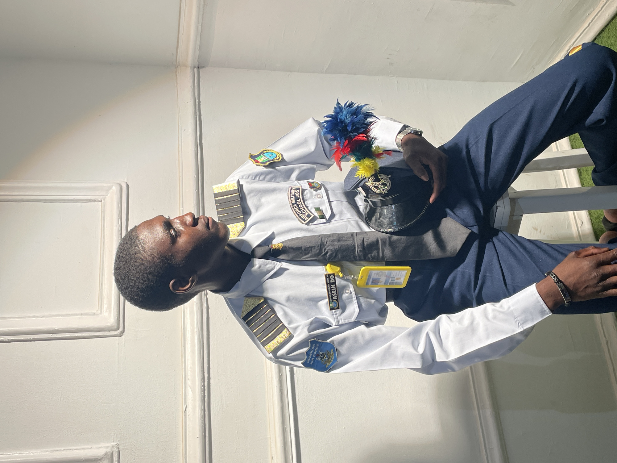 A young man in an Air Force uniform is reclining indoors against a paneled wall. He is holding a hat adorned with colored plumes. His uniform includes gold-striped epaulettes and patches indicating "Nigerian Air Force" and "Air Force School Jos". He is wearing a wristwatch and a bracelet. A name tag with the inscription "ASEFIN DO" is visible. The scene is brightly lit. There is no specific indication that this is in Barakin Akawo, Nigeria.