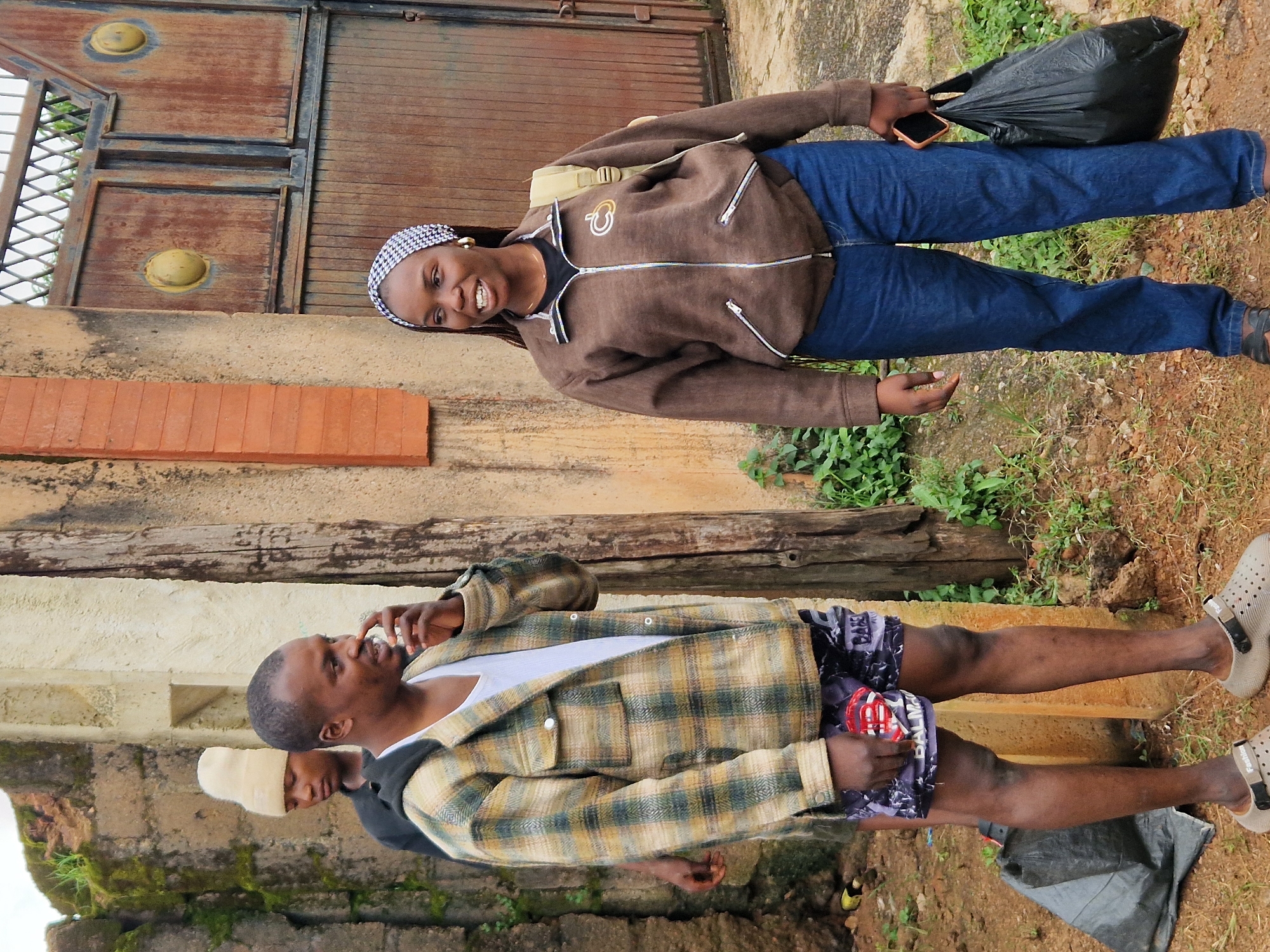 In Jos, Nigeria, two individuals stand outdoors near a textured, light-colored wall. The person on the left, a male, wears a plaid jacket over a white t-shirt and patterned shorts. He holds a mobile phone to his ear. To his right, a female wears a brown zippered jacket, a headscarf, and blue jeans. She carries a black plastic bag in her right hand. Behind them, a large, rusted metal gate is visible. The ground is a mix of dirt and scattered vegetation.