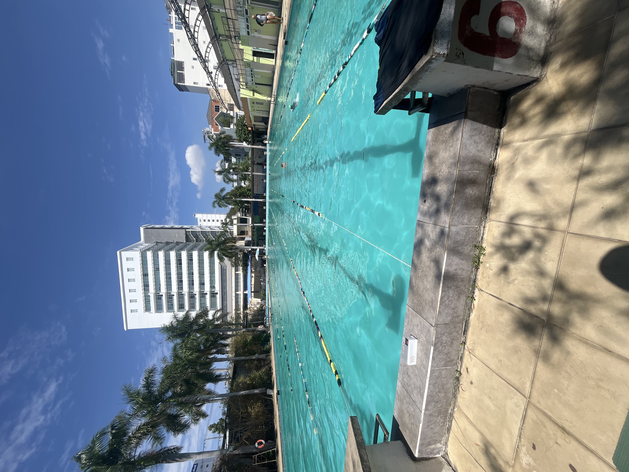 The image shows a swimming pool with lane dividers, adjacent to buildings, including a tall white building. The setting is outdoors under a blue sky with scattered clouds. There are palm trees near the pool and buildings. A diving block with the number "6" is visible at the edge of the pool. People appear to be swimming in the pool. The location is Nha Trang Ward, Vietnam.