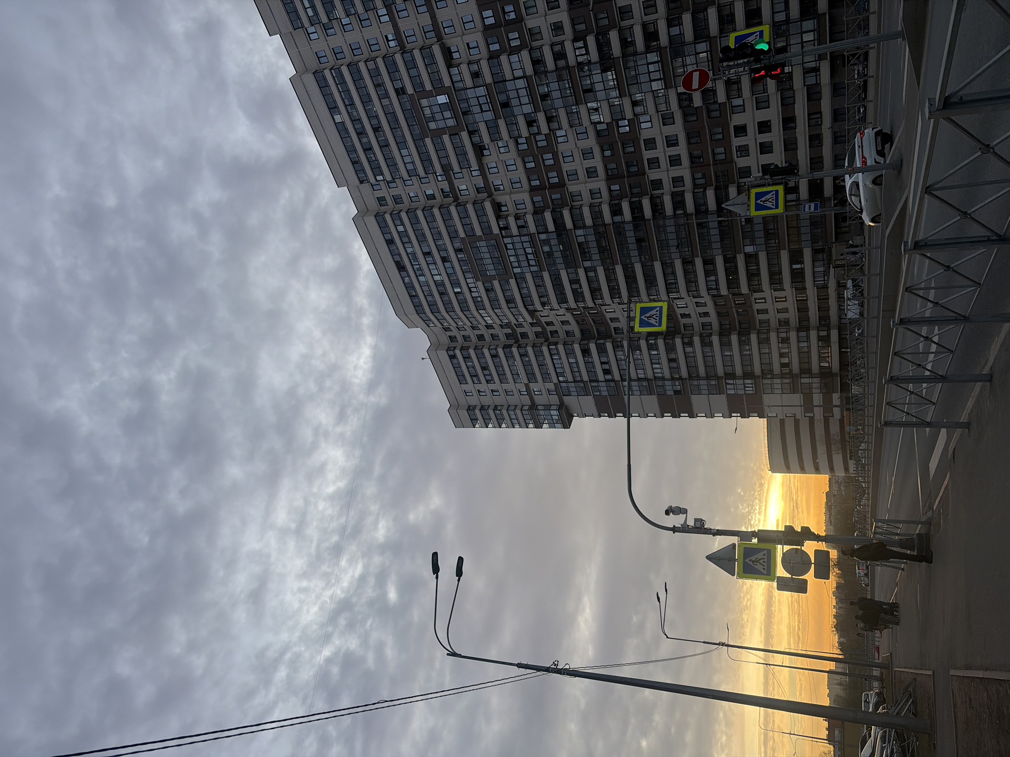 This image depicts a street scene at dusk in Saint Petersburg, Russia. A tall residential building dominates the left side of the frame, its facade marked by rows of windows. The sky above is filled with gray clouds. The right side of the image shows a street intersection with traffic lights displaying both red and green signals. Pedestrian crossing signs are visible near the traffic lights. A few individuals are standing on the sidewalk near the crossing. Street lamps are positioned along the street. The sunset glows in the distance, creating a bright orange horizon. A white car is parked near the traffic lights. Metal safety barriers line the edge of the sidewalk. Overhead power lines are present in the lower portion of the frame.