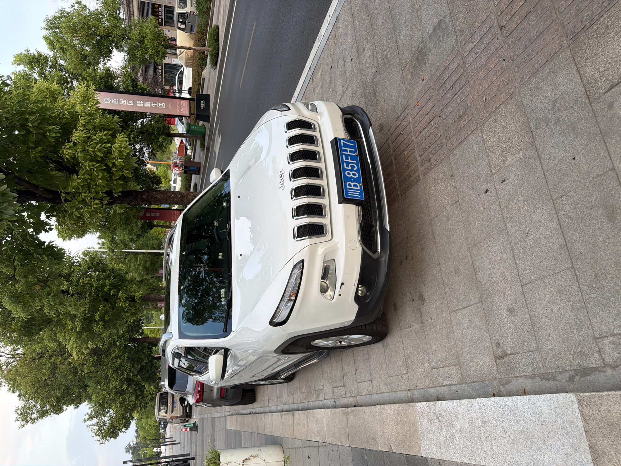 A white Jeep Cherokee is parked on a sidewalk in the city of Jiangjin, China. The vehicle is facing right, with its front end slightly angled towards the viewer. The Jeep has a license plate that reads "JWB-85FH7". In the background, a dark-colored SUV and a silver minivan are also parked on the street. Mature trees line the sidewalk, casting shadows on the ground. A vertical banner with Chinese characters stands near the right edge of the image. The sidewalk is paved with grey and pinkish granite tiles, and a tactile paving strip is visible on the right side. The street is asphalt. The sky is overcast.