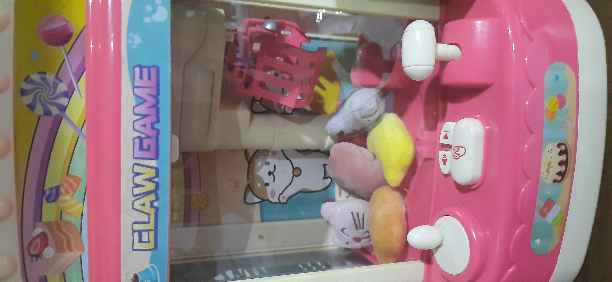 The image shows a close-up of a pink toy claw machine. The machine has a clear plastic front, revealing a colorful interior decorated with cartoon images of cats and candies. Inside, there are several plush toys, including a white cat-shaped toy, a pink oval toy, and a yellow star-shaped toy. The controls of the machine are visible at the bottom, featuring a joystick and buttons. The machine is positioned in an indoor environment, likely a home or a toy store, and it is not possible to determine its specific location in Cisauk, Indonesia from the provided image.