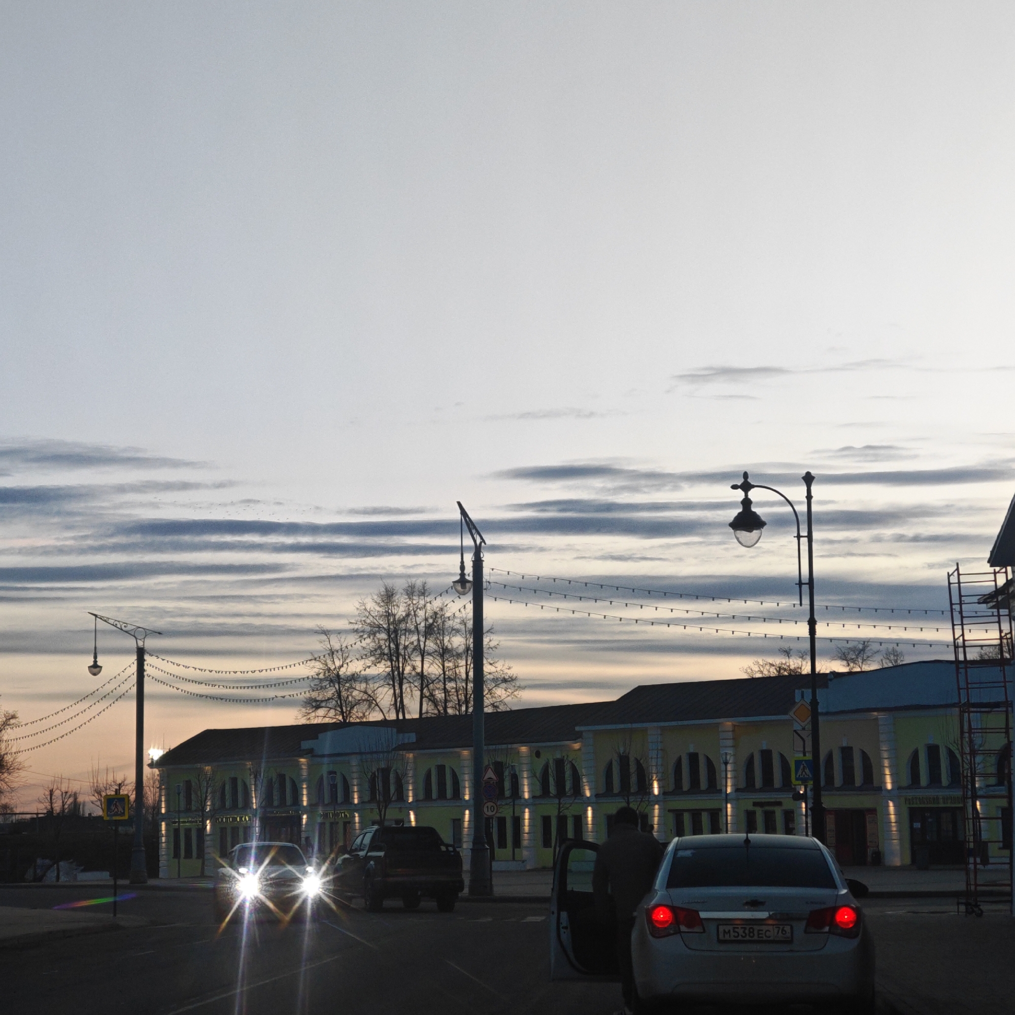 The image shows a street scene in what appears to be the city of Rostov, Russia. The time of day seems to be dusk or early evening, with light illuminating the sky. The sky has a pattern of horizontal, textured clouds. In the middle ground, a two-story building with arched windows runs along the street. The building is painted in yellow and white. Decorative lights hang from lampposts along the street. In the foreground, there are several cars, one of which has an open door and a person is partially visible near it. There are other vehicles, including one with headlights on, further down the street. The street appears to be a paved road. There is scaffolding on the right side of the image near the building. Overall, the scene captures an urban environment with a mix of architectural elements and street-level activity.