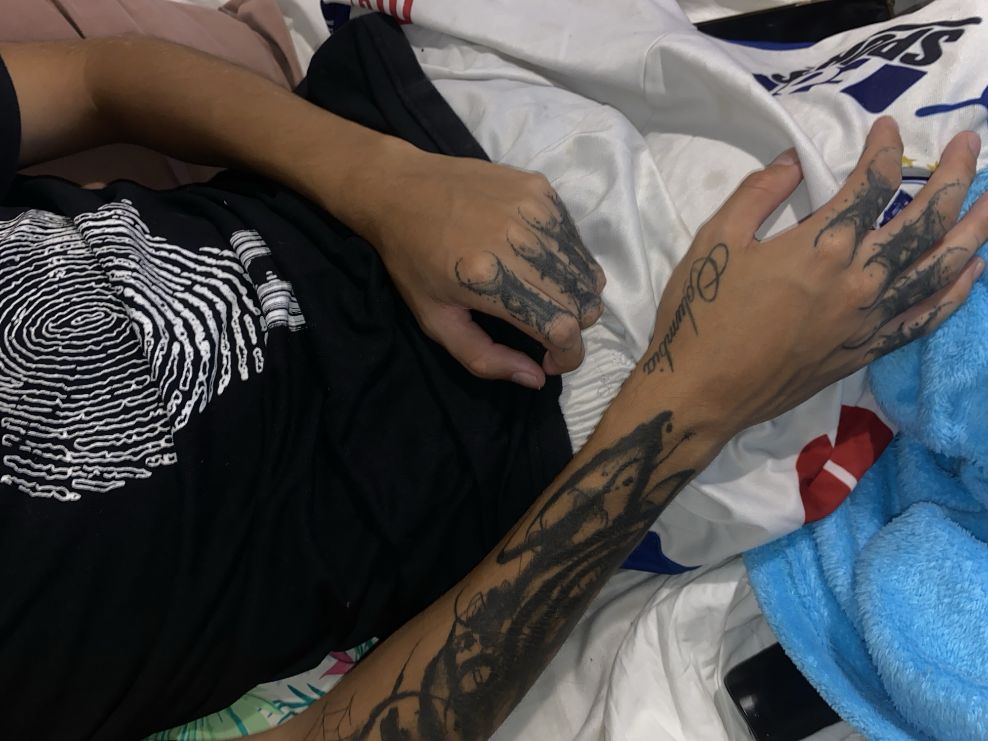 The image shows a person lying on a bed. The person is wearing a black t-shirt with a white fingerprint design. Both arms are visible and have multiple tattoos. The right arm has a tattoo that says "Columbia" and another tattoo of a design that extends from the hand to the forearm. The left hand also has black tattoos on the fingers and back of the hand. The bedding is white with some areas featuring red, blue, and white patterns. A blue towel is also present on the bed. There is no information in the image to suggest the location is in Buenos Aires, Argentina.