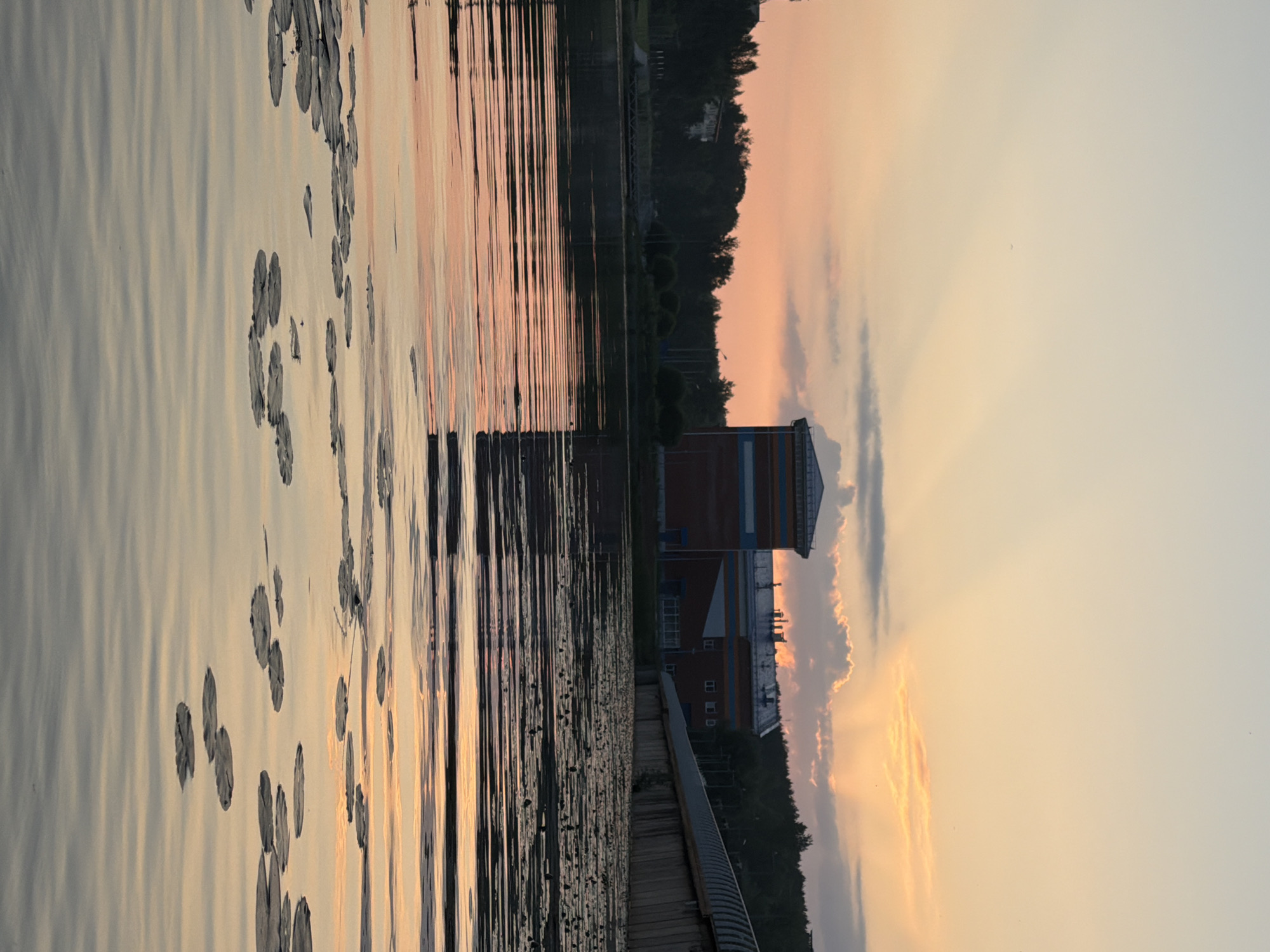 A serene sunset casts a warm glow over a body of water dotted with lily pads. In the background, a modern building with a brick facade and blue accents stands against a backdrop of trees. A curved concrete structure lines the water's edge. The sky is painted with hues of orange and pink, with rays of light piercing through scattered clouds. The water reflects the colors of the sky, creating a tranquil and picturesque scene.