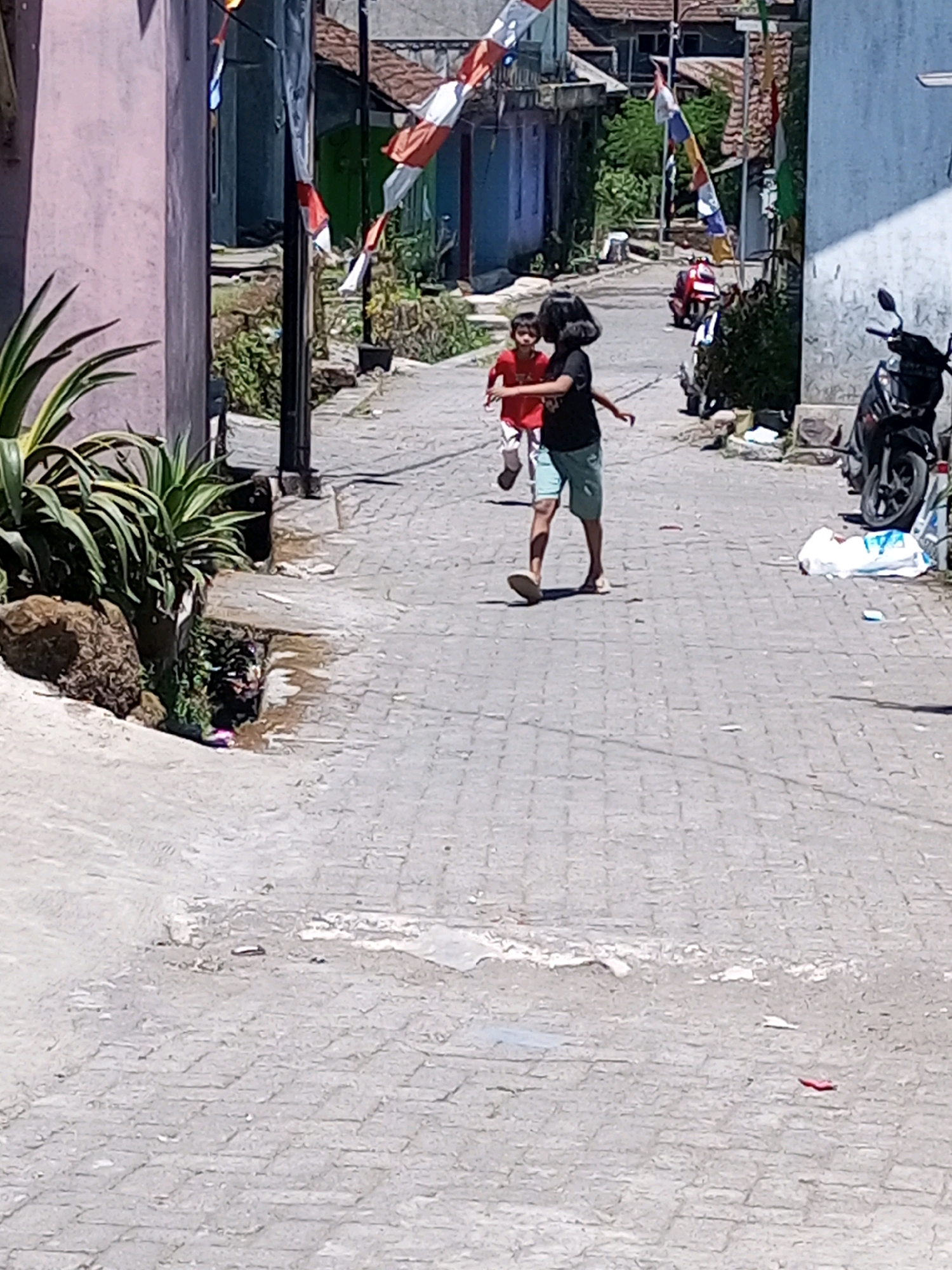 Two children are running down a paved village street in Andongsili, Indonesia. The child in the foreground, wearing a black t-shirt and blue shorts, is kicking their left leg forward as they run. The second child, further back and to the left, wears a red shirt and white shorts and is also running. They are on a street with cobblestone paving, flanked by buildings on either side. Flags, some red and white, are hung along the street, suggesting a possible celebration or national holiday. A motorcycle is parked on the right side of the street. Green vegetation is visible along the sides of the street and in front of some of the buildings. The scene appears to be taking place during daylight hours, with shadows indicating the sun is overhead.
