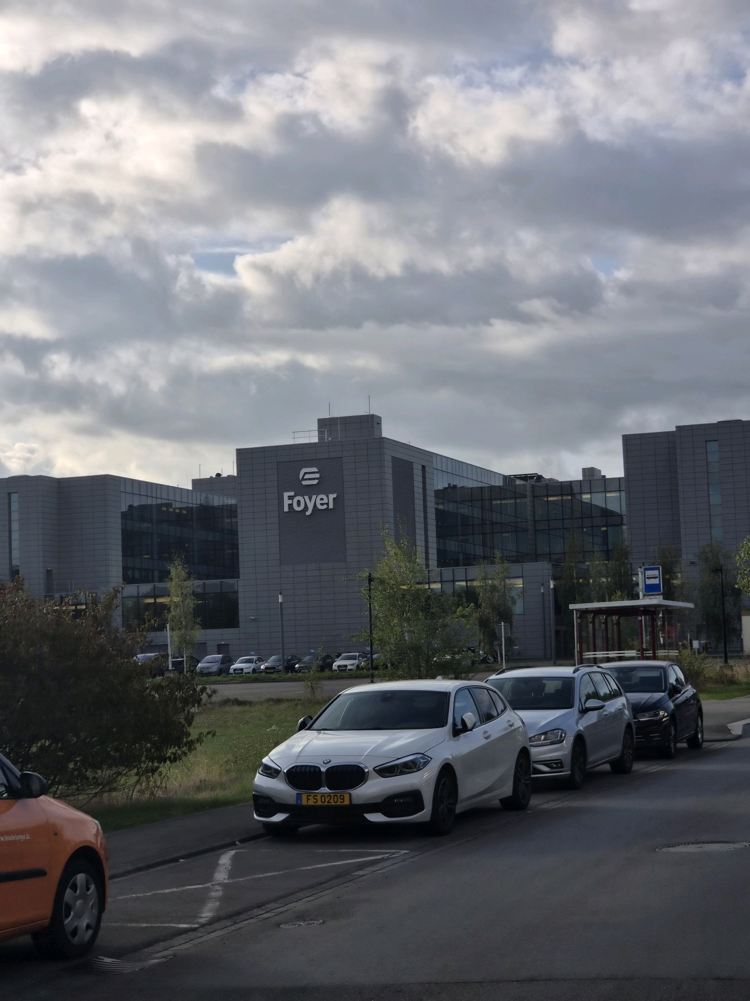 The image shows a modern office building with the "Foyer" logo visible on its facade, located in Leudelange, Luxembourg. The building has a gray exterior and large glass windows reflecting the cloudy sky. In front of the building, several cars are parked in a row. Among them, a white BMW with license plate "FS 0209" is prominently featured. An orange car is partially visible in the foreground. A bus stop shelter with a blue sign is also present near the building. The overall environment appears to be an urban or business park setting, under a heavily overcast sky.