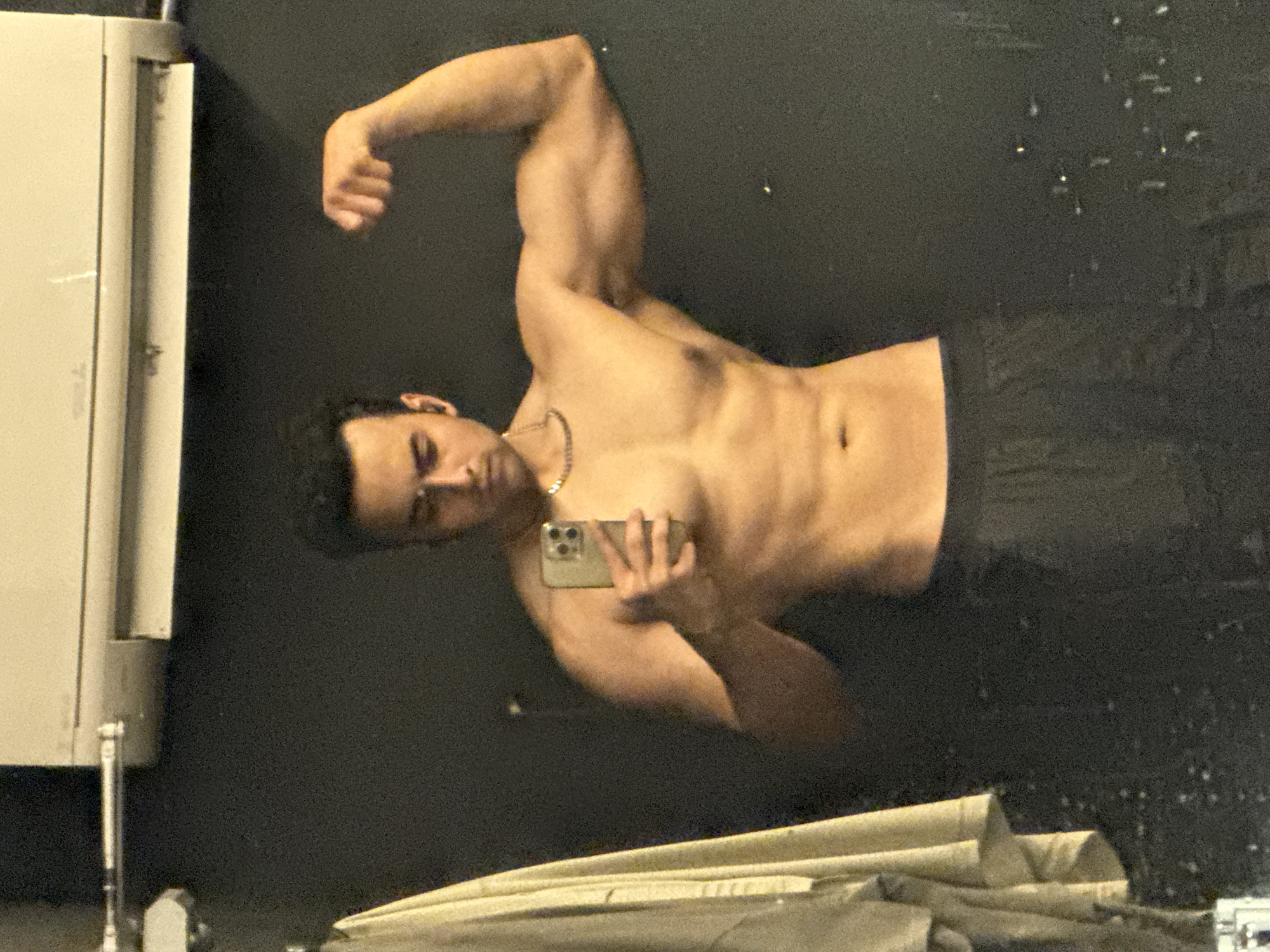 A young man, appearing to be in his early twenties, is captured in a mirror selfie taken indoors. He has dark, curly hair and is shirtless, showcasing a muscular physique. He is wearing black athletic shorts and a silver chain necklace. His right arm is flexed, highlighting his bicep. He is holding a smartphone in his left hand, positioned to take the picture. The background is a dark, textured wall, with what appears to be an air conditioning unit mounted above. A light-colored garment is hanging on the left side of the frame, partially obscuring the wall. The image quality is somewhat grainy, suggesting it might have been taken in low light conditions. There is no discernible information within the image that places the location in Kolkata, India.